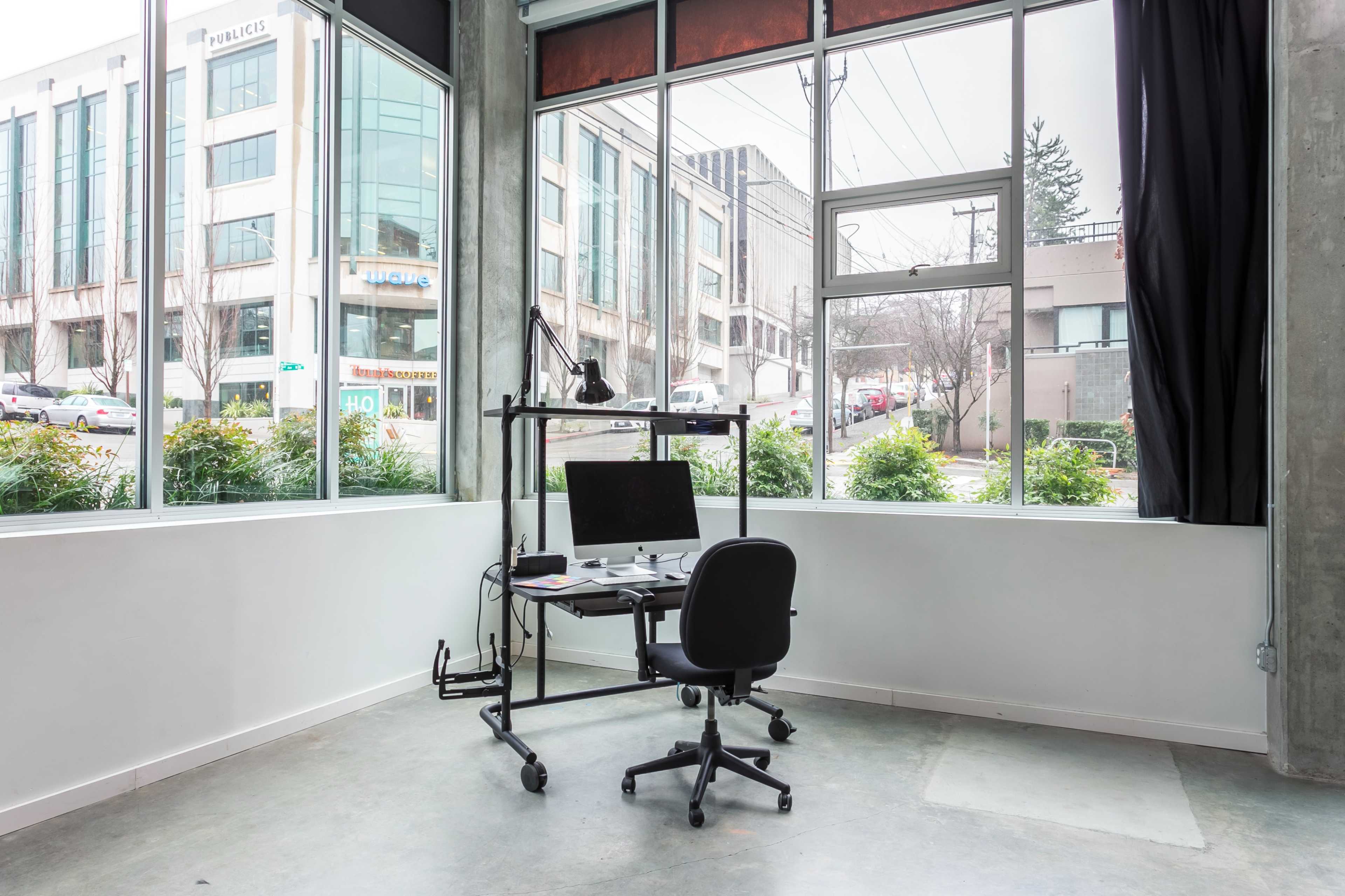 The image shows a modern office space with a standing desk and chair positioned near large windows overlooking a city street.