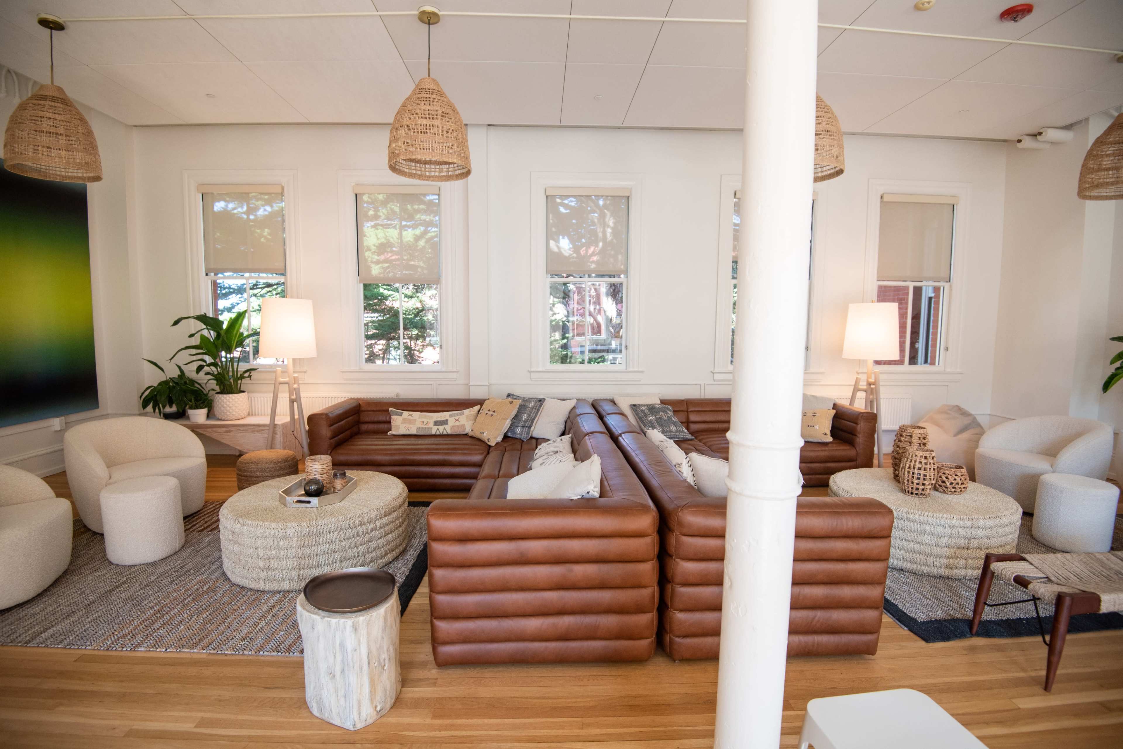 A modern living room with a large leather sectional sofa, light-colored chairs, a coffee table, and various decorative plants.