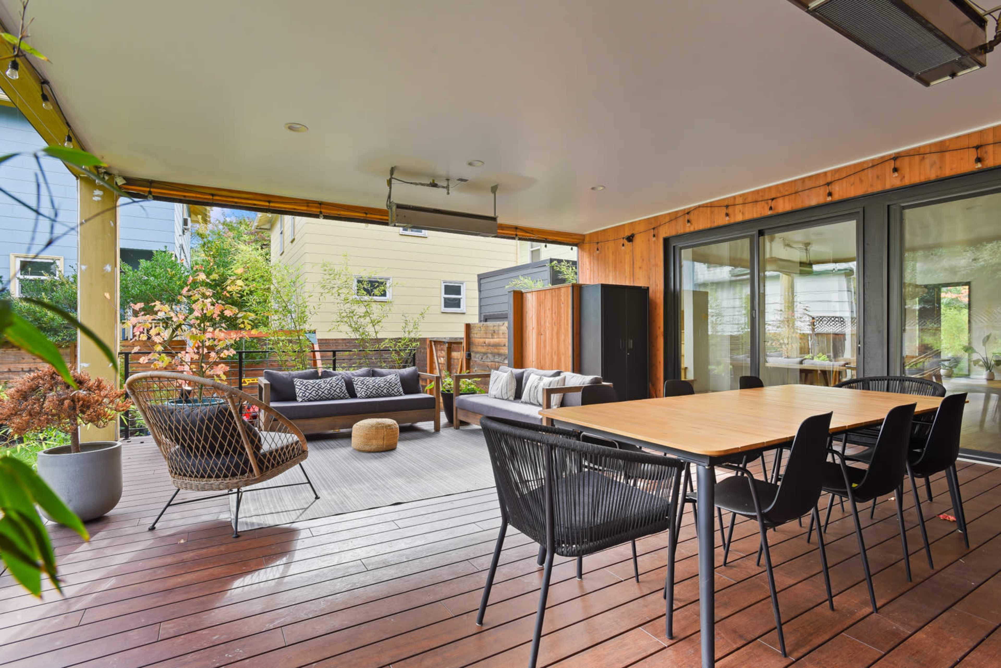 The image shows a covered outdoor patio featuring a dining table, black chairs, a seating area with a sofa, and potted plants.