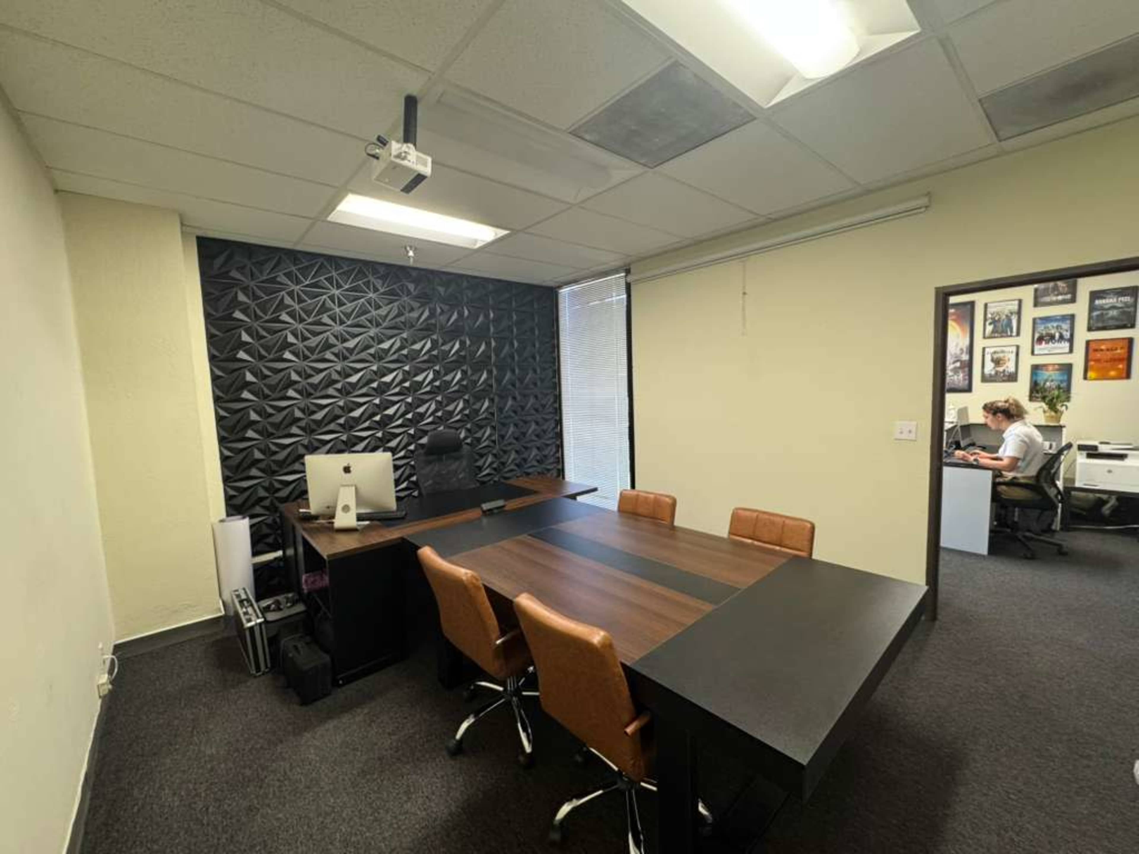 The image shows a modern office space featuring a wooden conference table surrounded by chairs, an ergonomic desk with a computer, and a textured black accent wall.