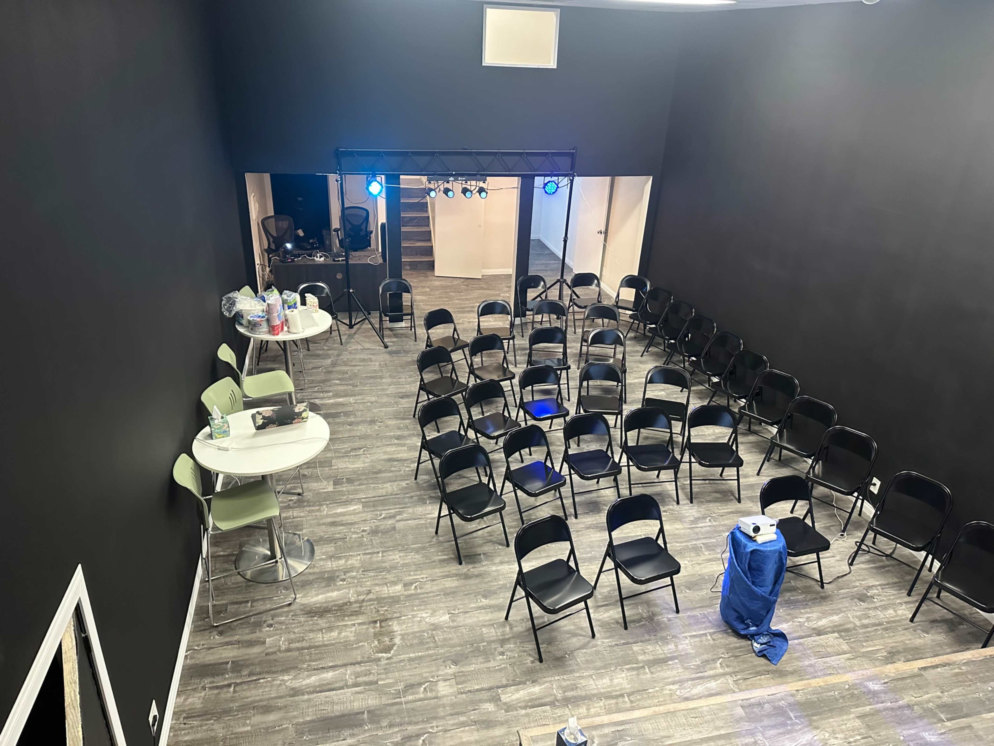 The image shows a spacious room set up with rows of black folding chairs facing a stage area, with a bar table and a few items on the floor.