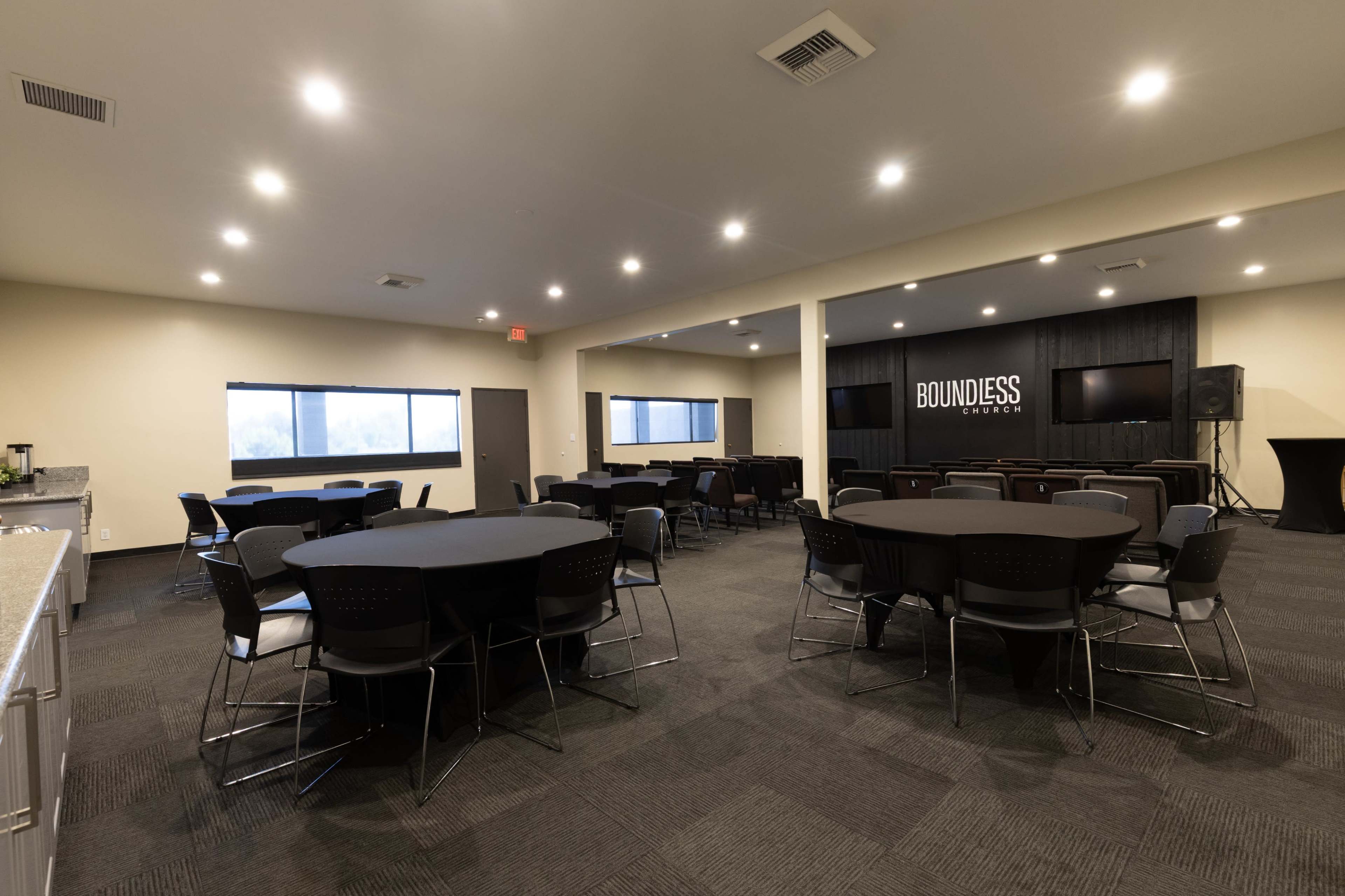 The image shows a modular event space with several round tables and chairs arranged in a well-lit room, featuring a kitchen area and a large screen on one wall.