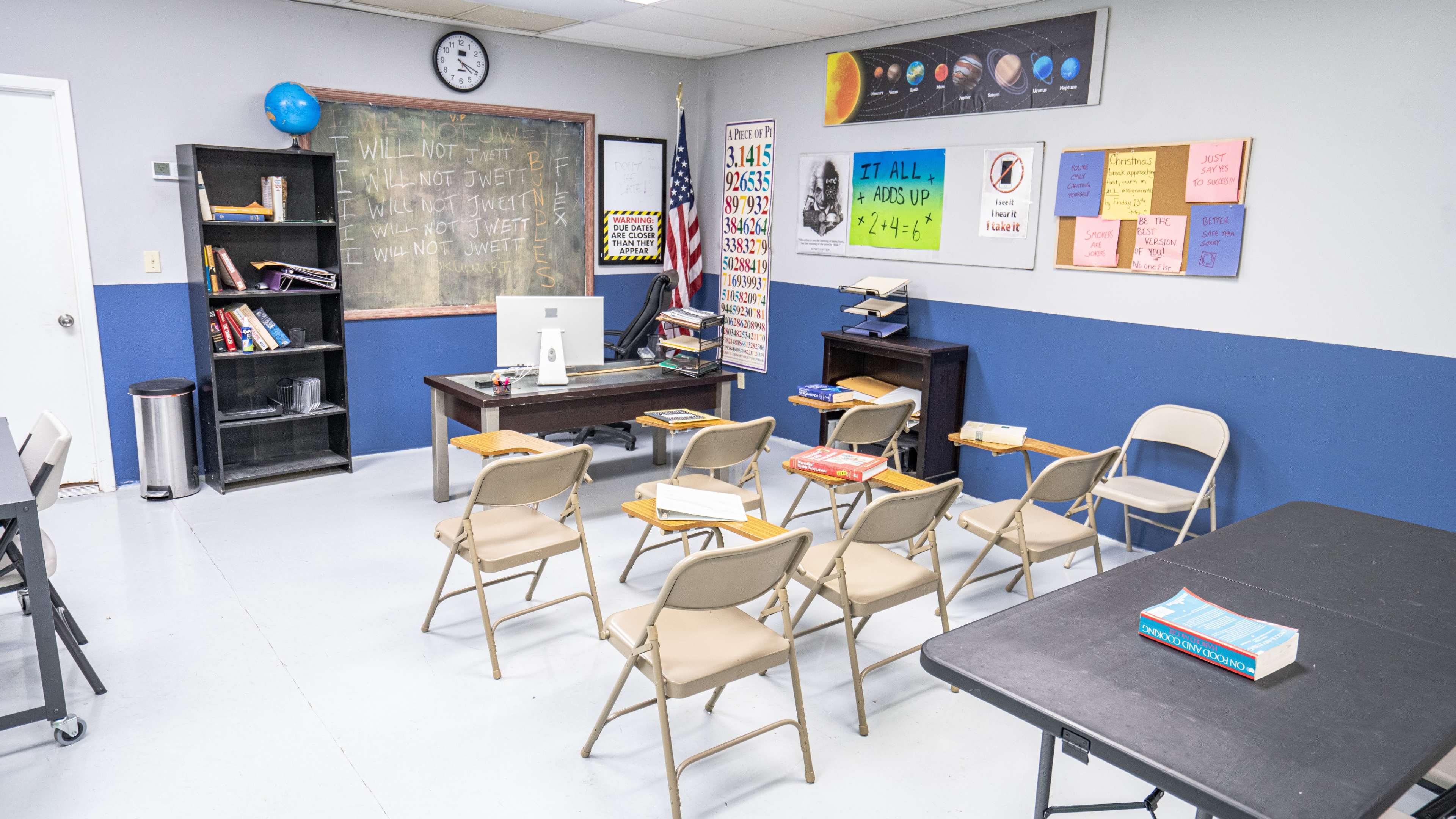 A classroom is set up with empty chairs facing a desk and a chalkboard, featuring educational posters and books scattered on the tables.