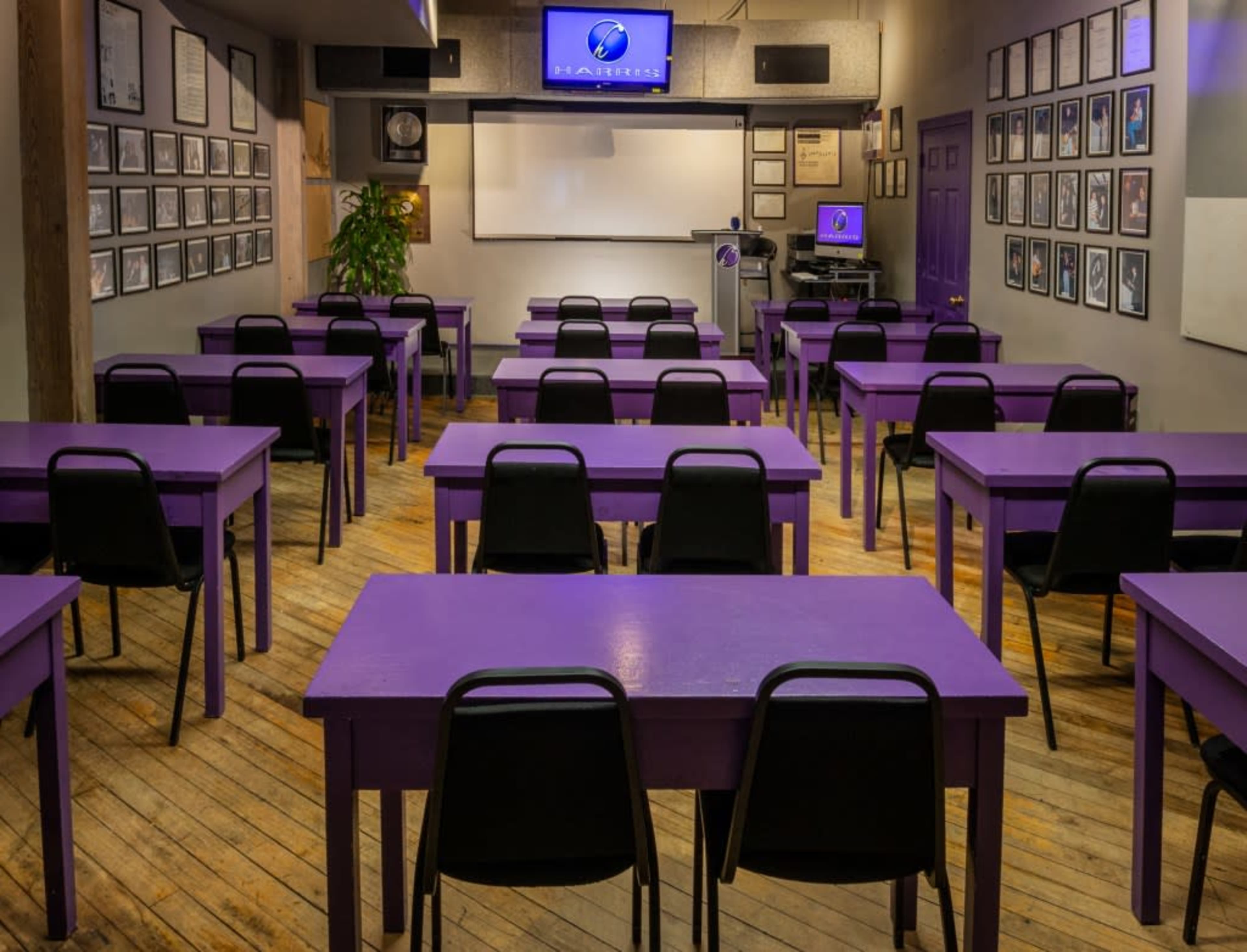 The image shows a classroom with purple tables arranged in rows and framed photographs adorning the walls.