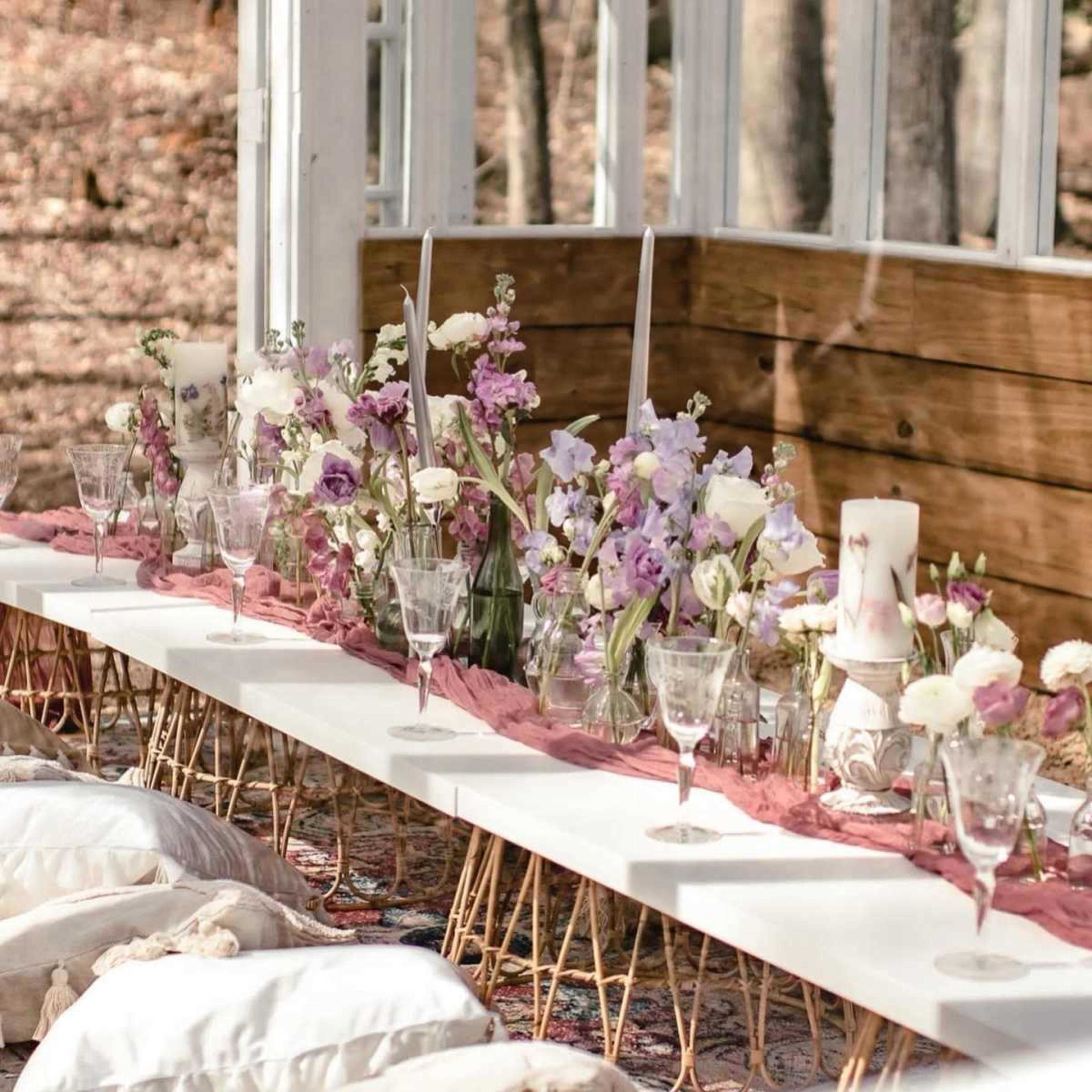 A long, white table is decorated with floral centerpieces and glassware, set up inside a tent-like structure surrounded by trees.