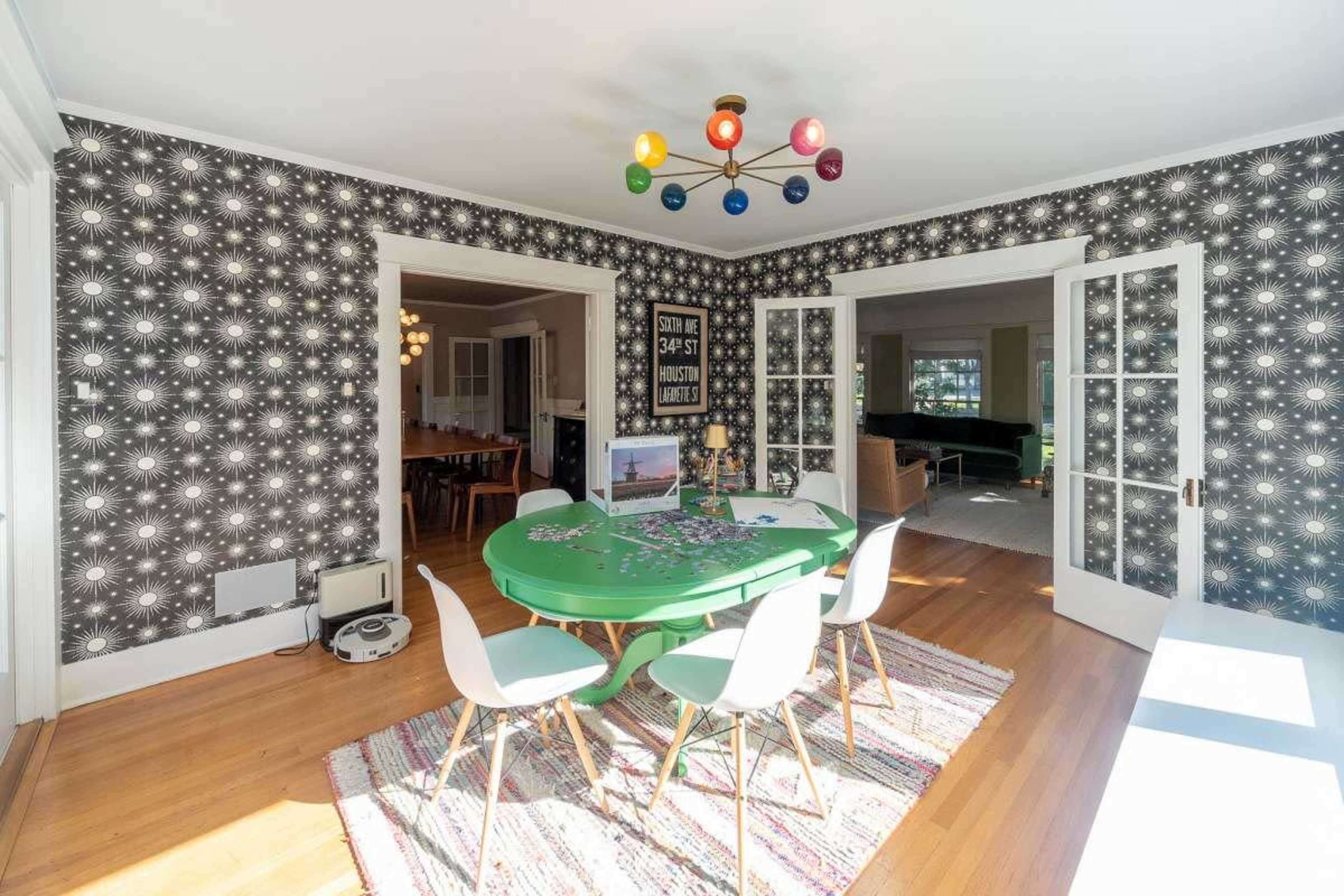 A green table with white chairs sits in a brightly lit room featuring patterned wallpaper and a colorful light fixture overhead.