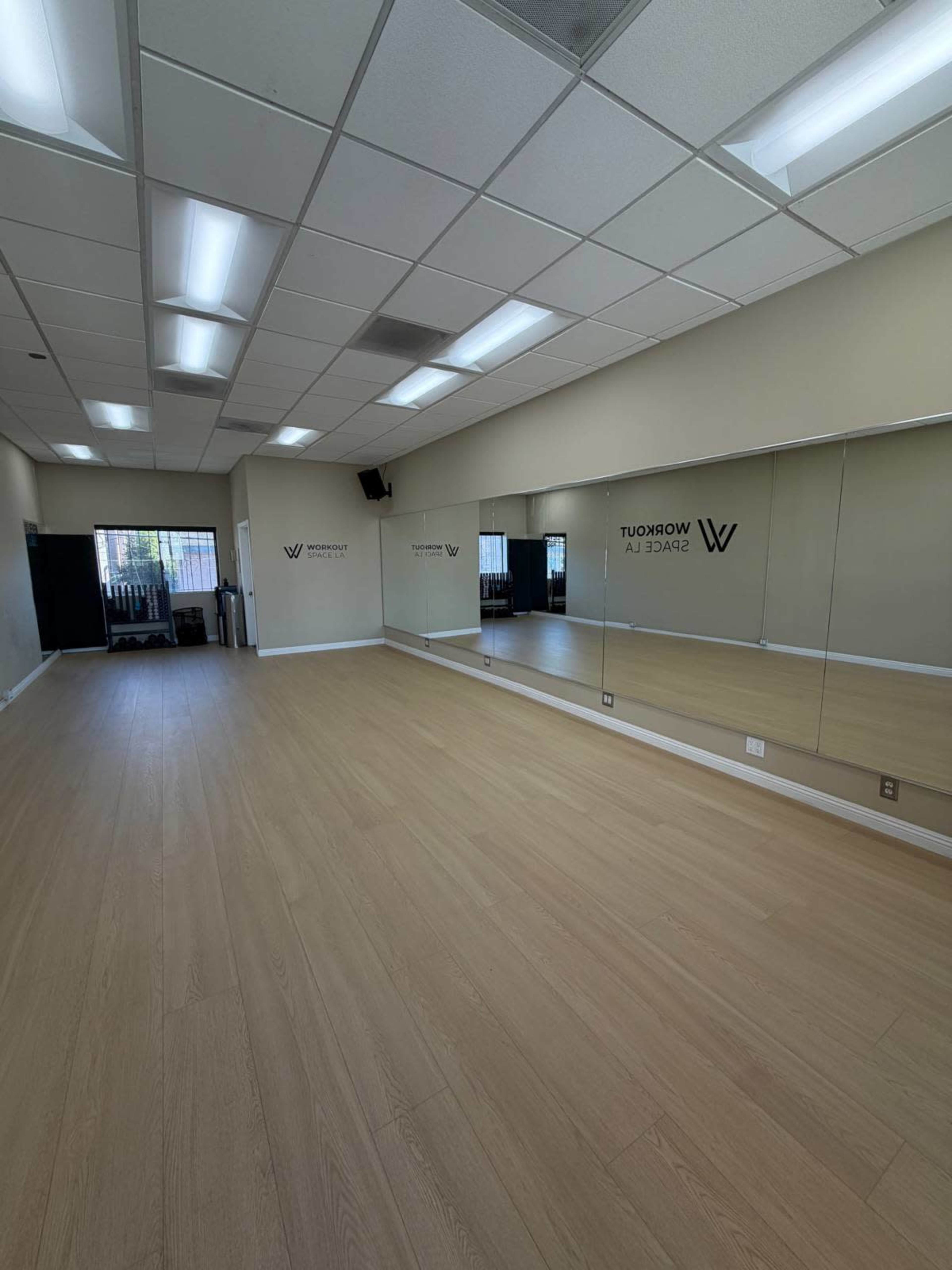 The image shows a spacious, empty room with wooden flooring and large mirrors on one wall, featuring the name "W" on the adjacent wall.