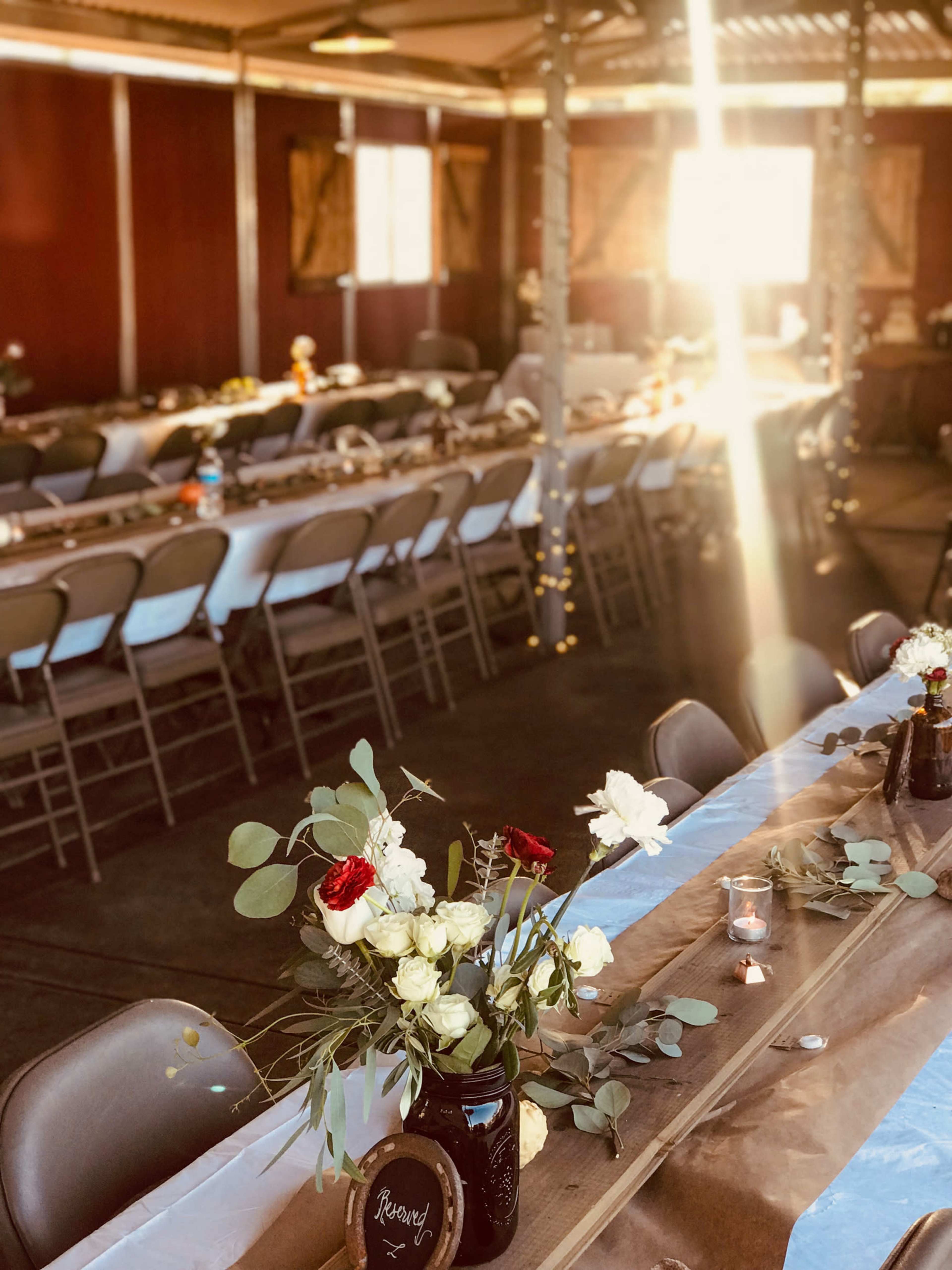 A well-decorated indoor venue features long tables set with floral centerpieces, chairs arranged neatly, and sunlight streaming through a window.
