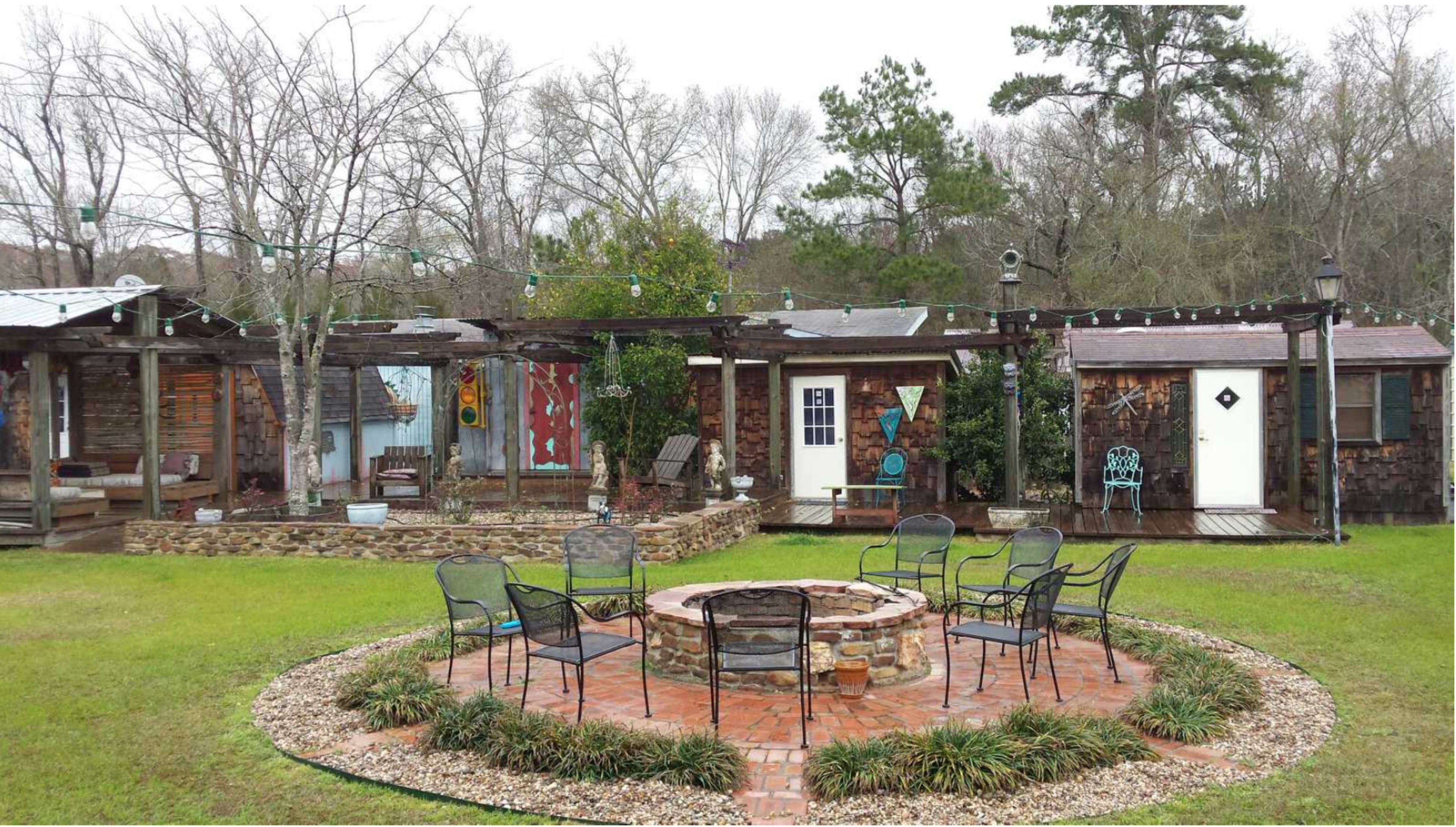 A circular stone fire pit surrounded by several metal chairs in a yard with wooden structures and trees in the background.