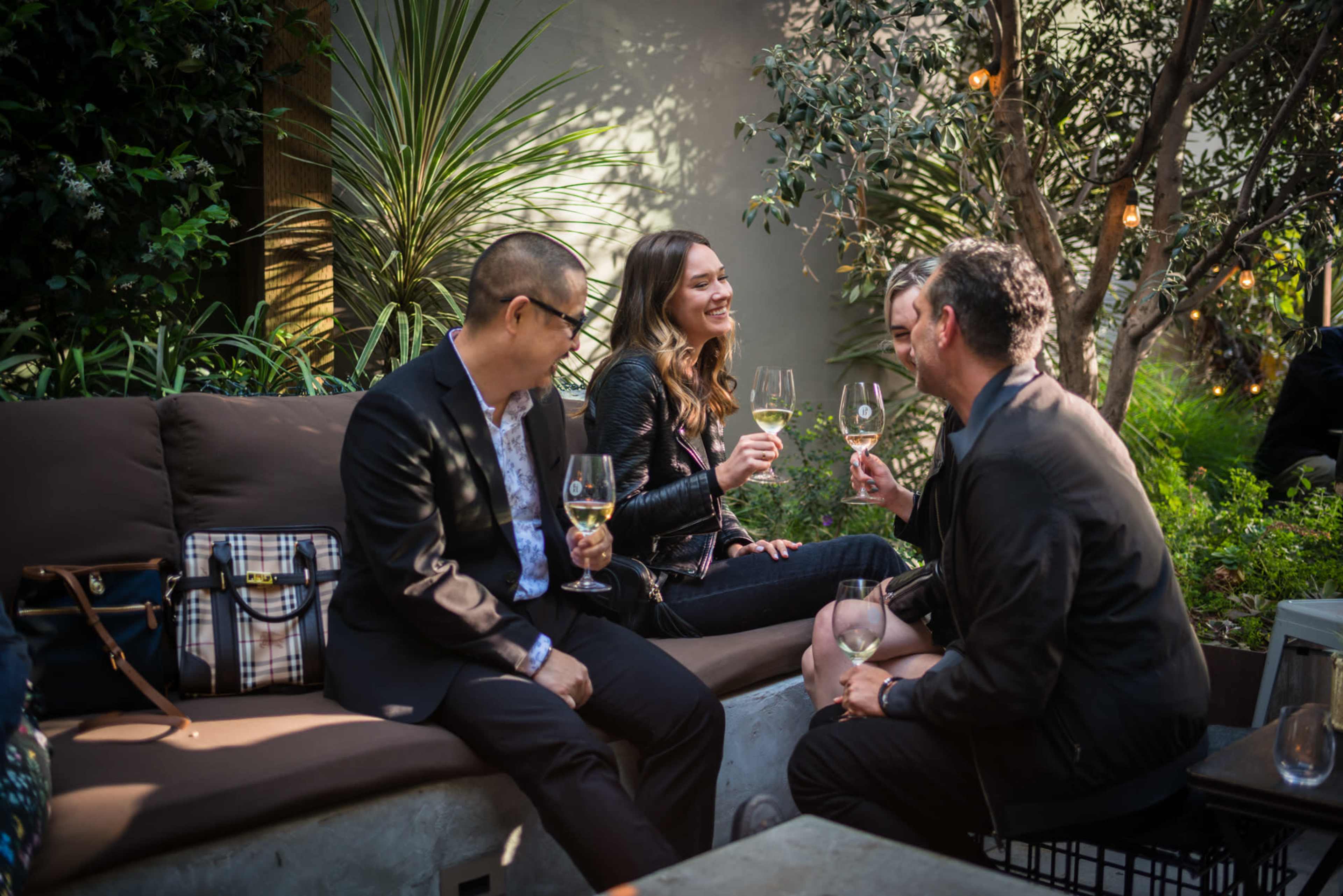 Four people sit together on a bench in a garden setting, enjoying drinks and engaging in conversation.