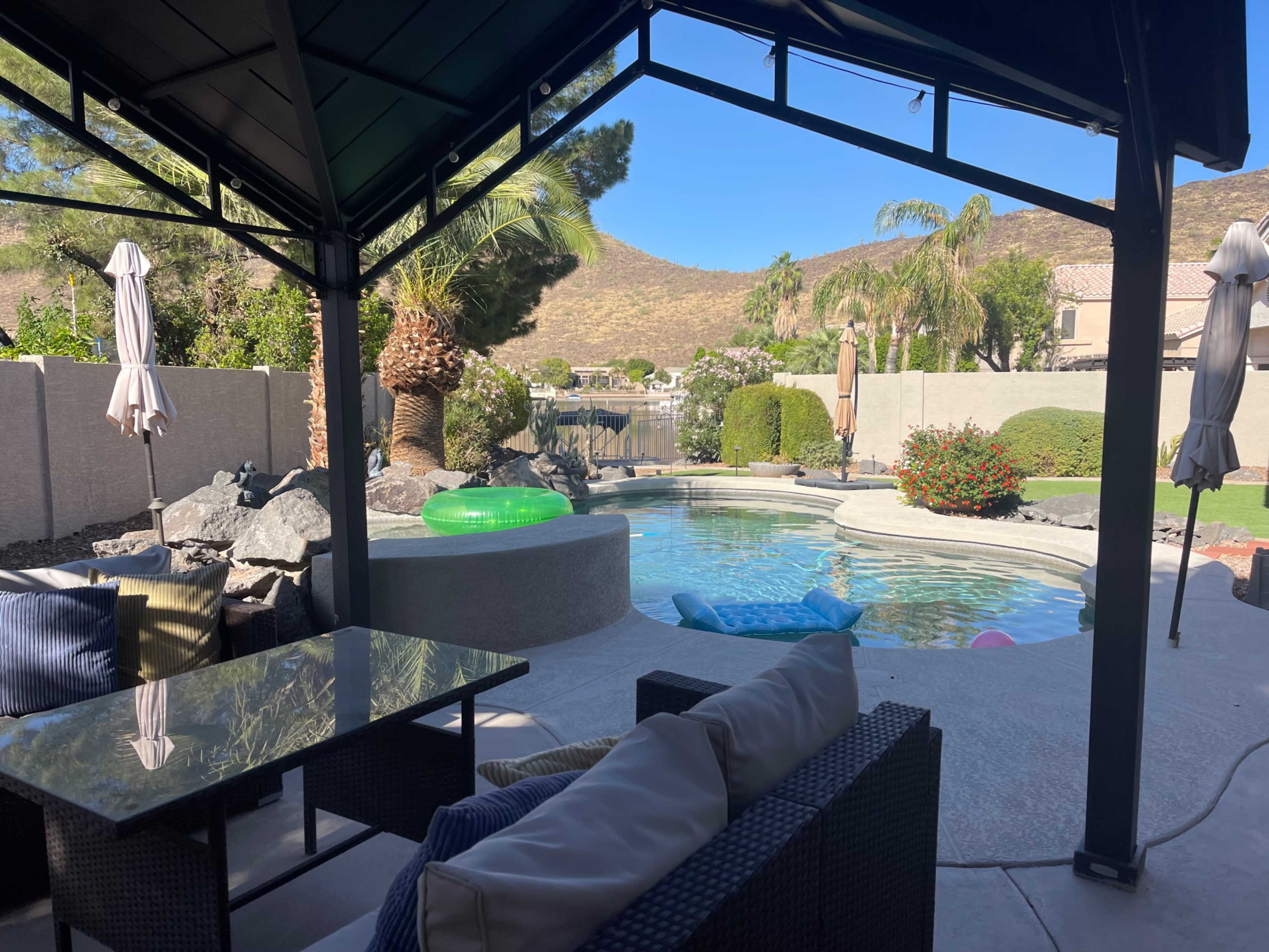 A shaded patio area overlooking a swimming pool surrounded by rocks, greenery, and a scenic view of a hillside.