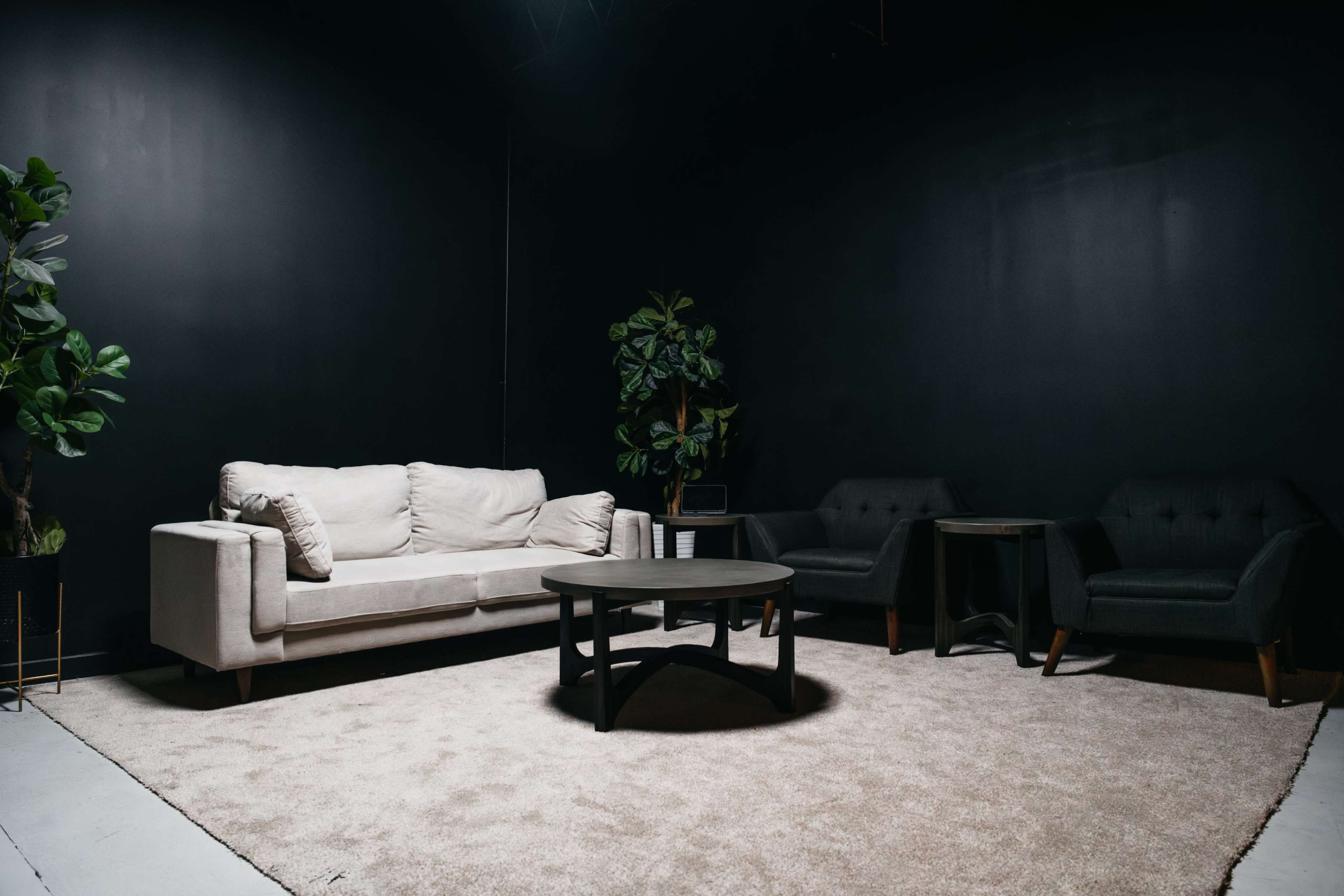 The image shows a minimalist lounge area with a light-colored sofa, two dark chairs, and a round table on a light carpet against a black wall.