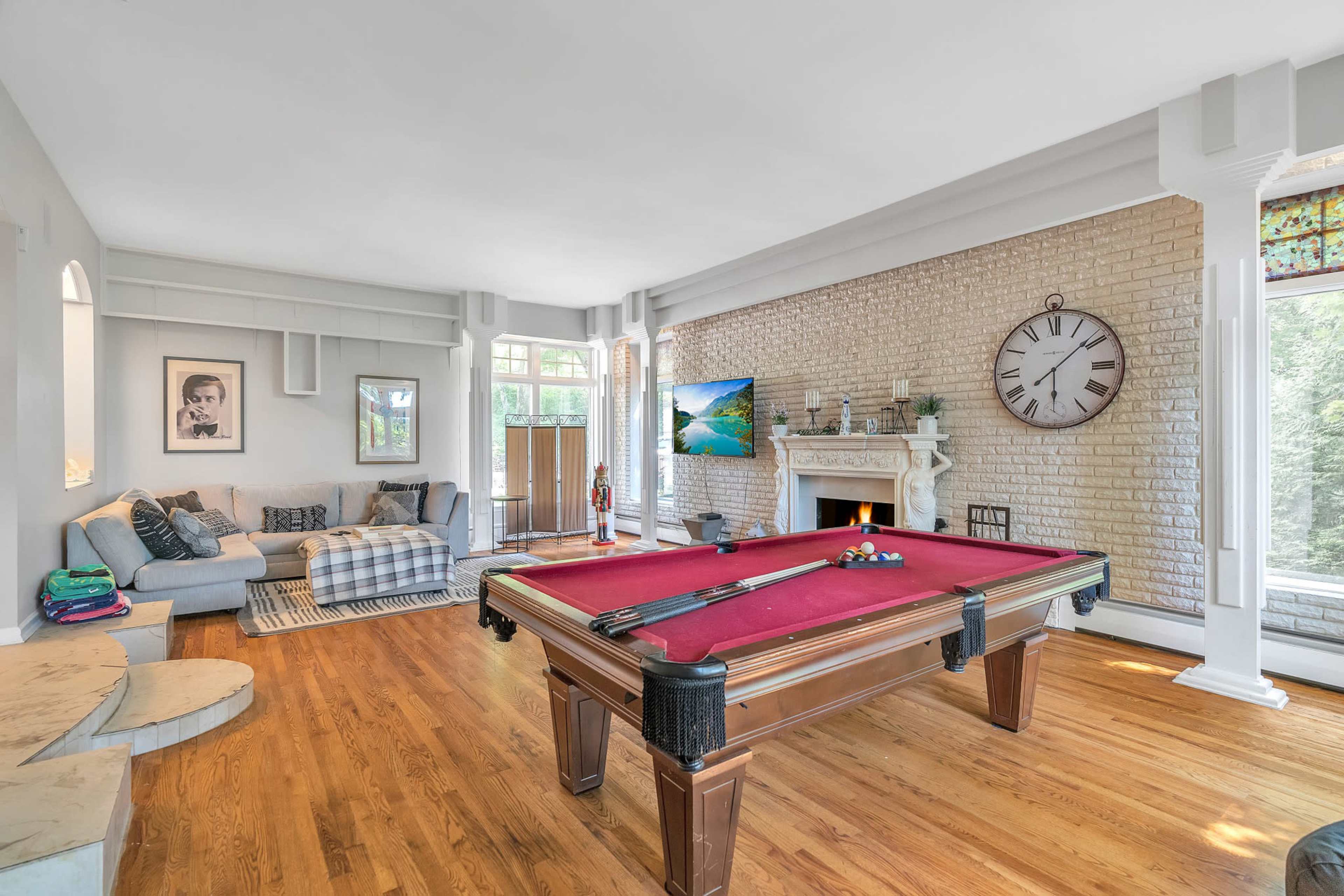 A spacious living room with a red pool table, a cozy seating area, and a large clock on the brick wall.