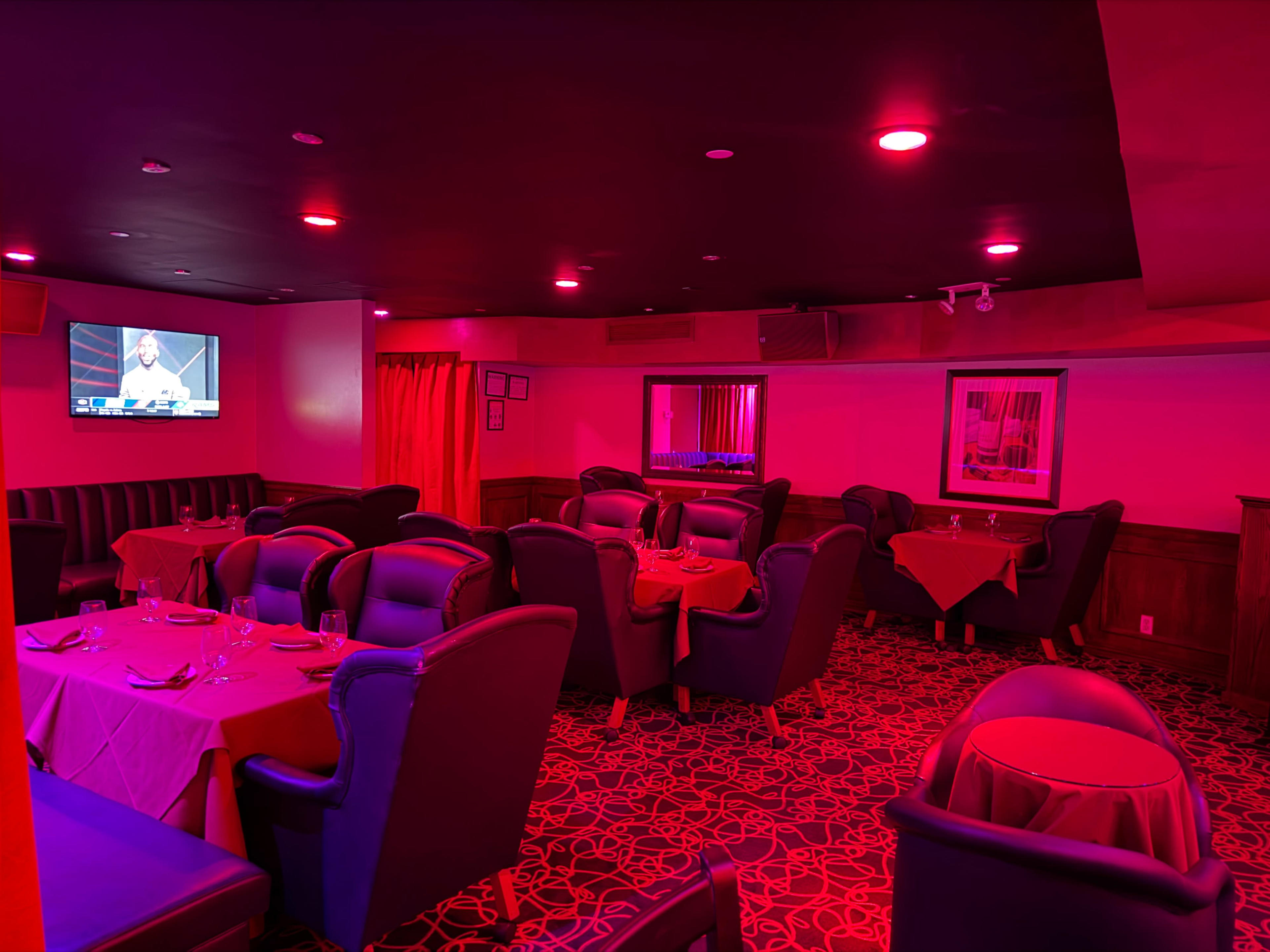 The image shows a dimly lit restaurant with red lighting, featuring tables set with white tablecloths and black leather chairs.