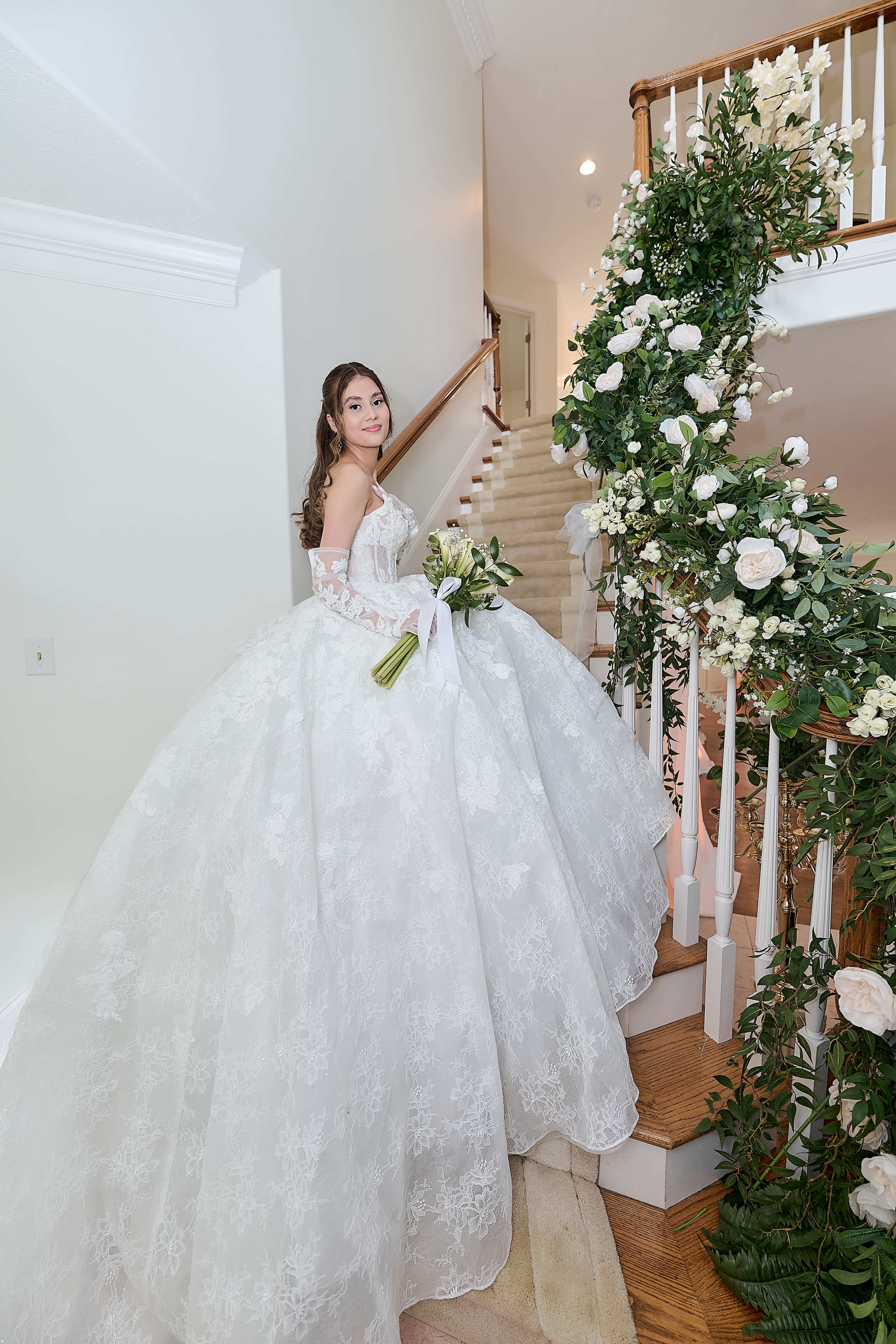 A bride in a luxurious white wedding dress stands on a staircase adorned with greenery and white flowers, holding a bouquet.