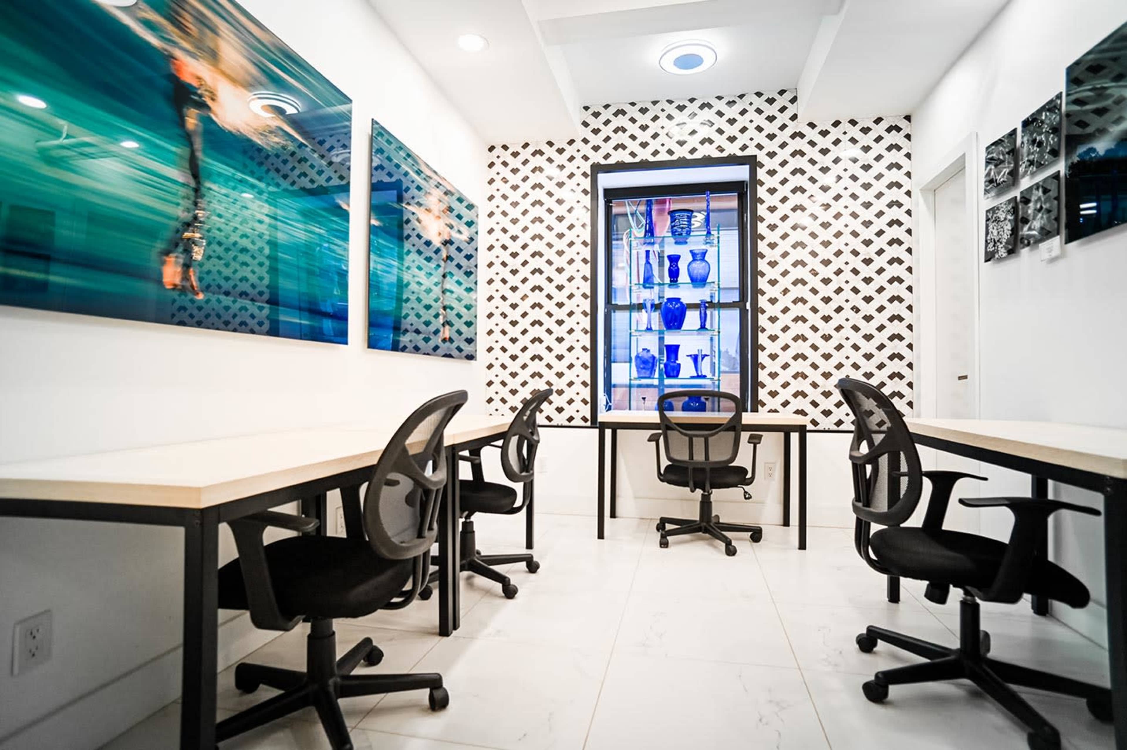 The image shows a modern workspace featuring two desks and chairs arranged in a bright room with artwork and a display cabinet in the background.