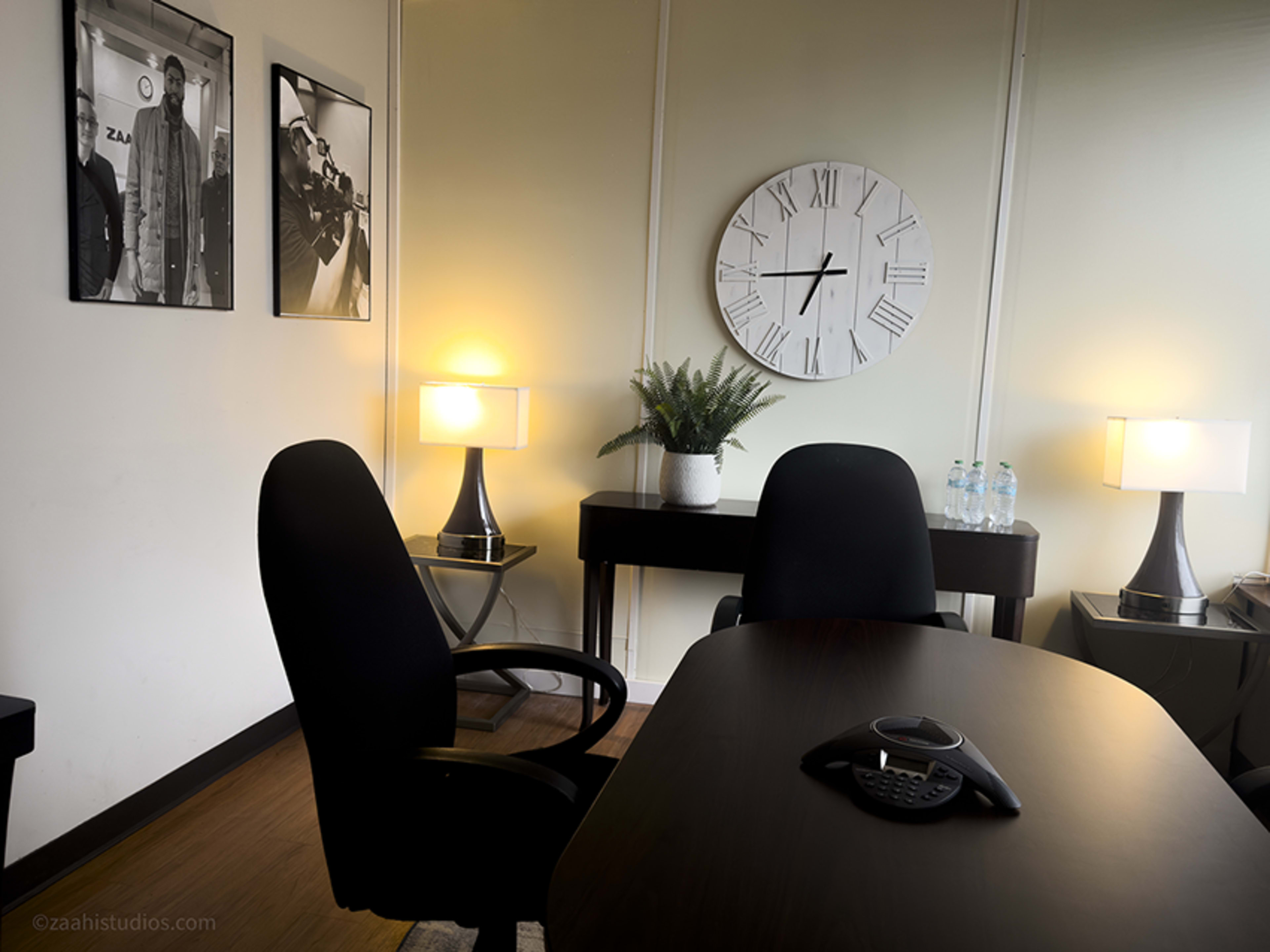 The image shows a modern office space with two black chairs facing a dark wooden desk, featuring a phone, a large wall clock, and two lamps on either side.