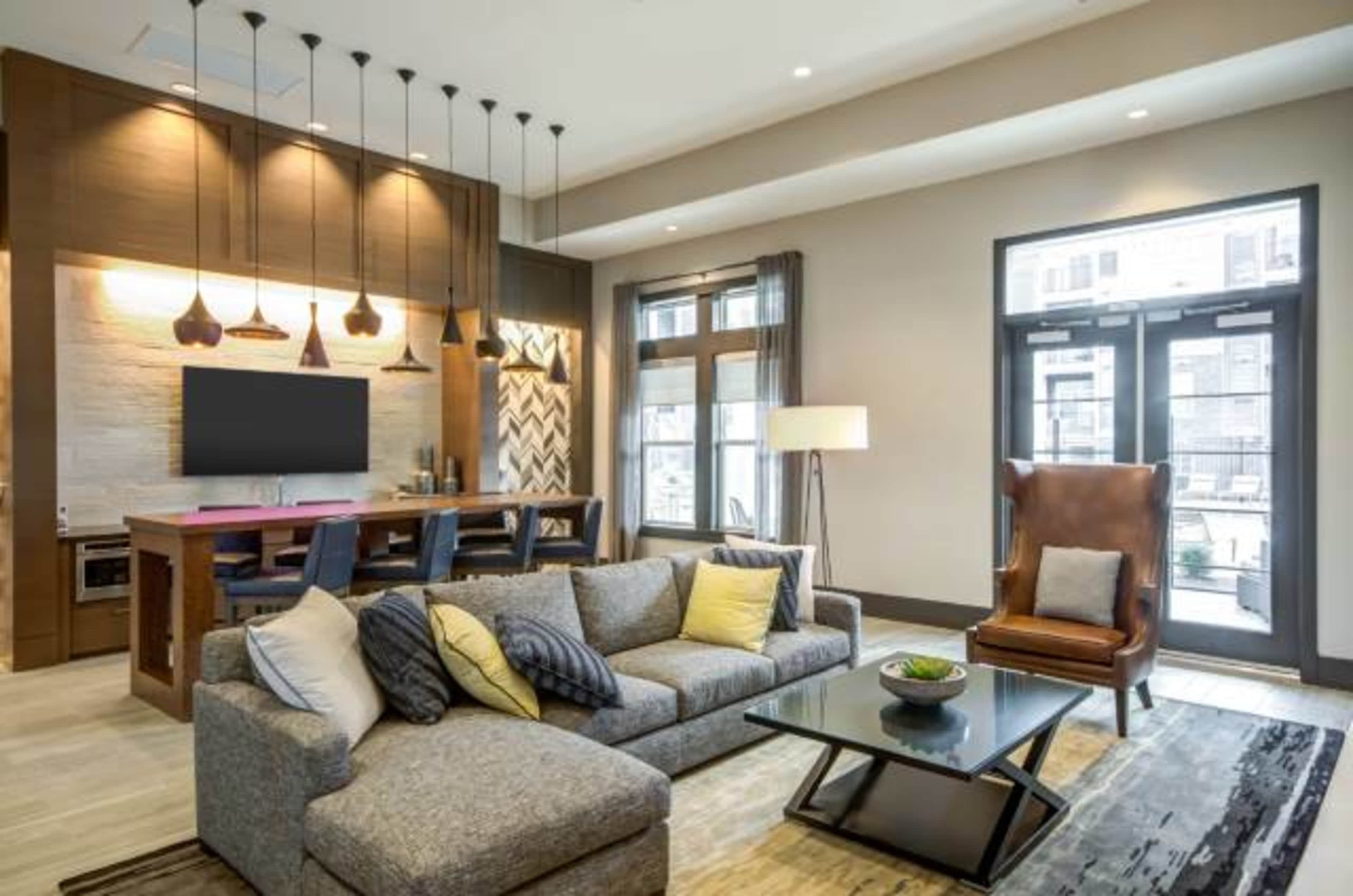 A modern living room with a gray sectional sofa, a black coffee table, a TV mounted on a wooden feature wall, and large windows leading to an outdoor area.