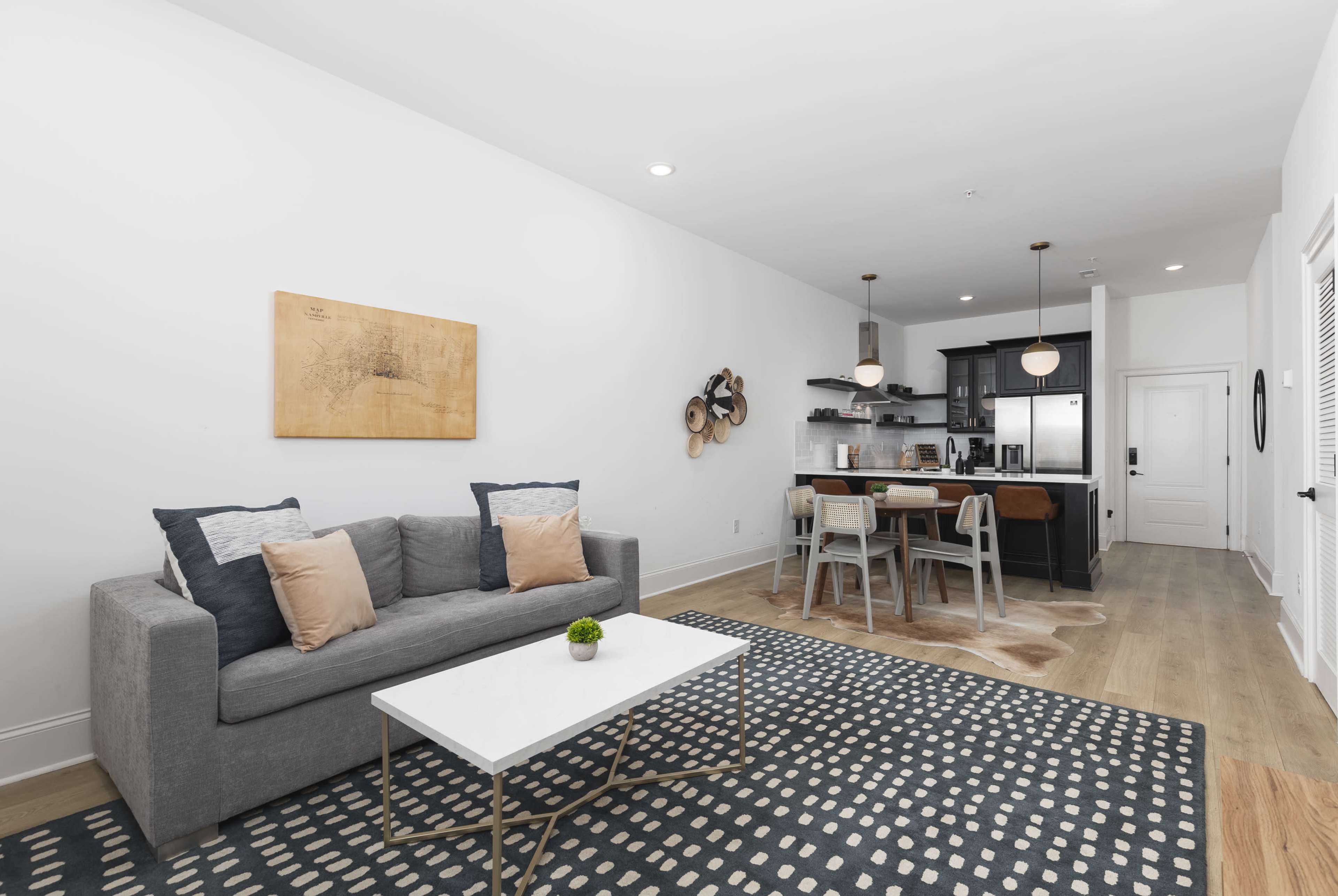 A modern living room features a gray sofa with decorative pillows, a white coffee table, and a kitchen area in the background with dark cabinetry and bar seating.