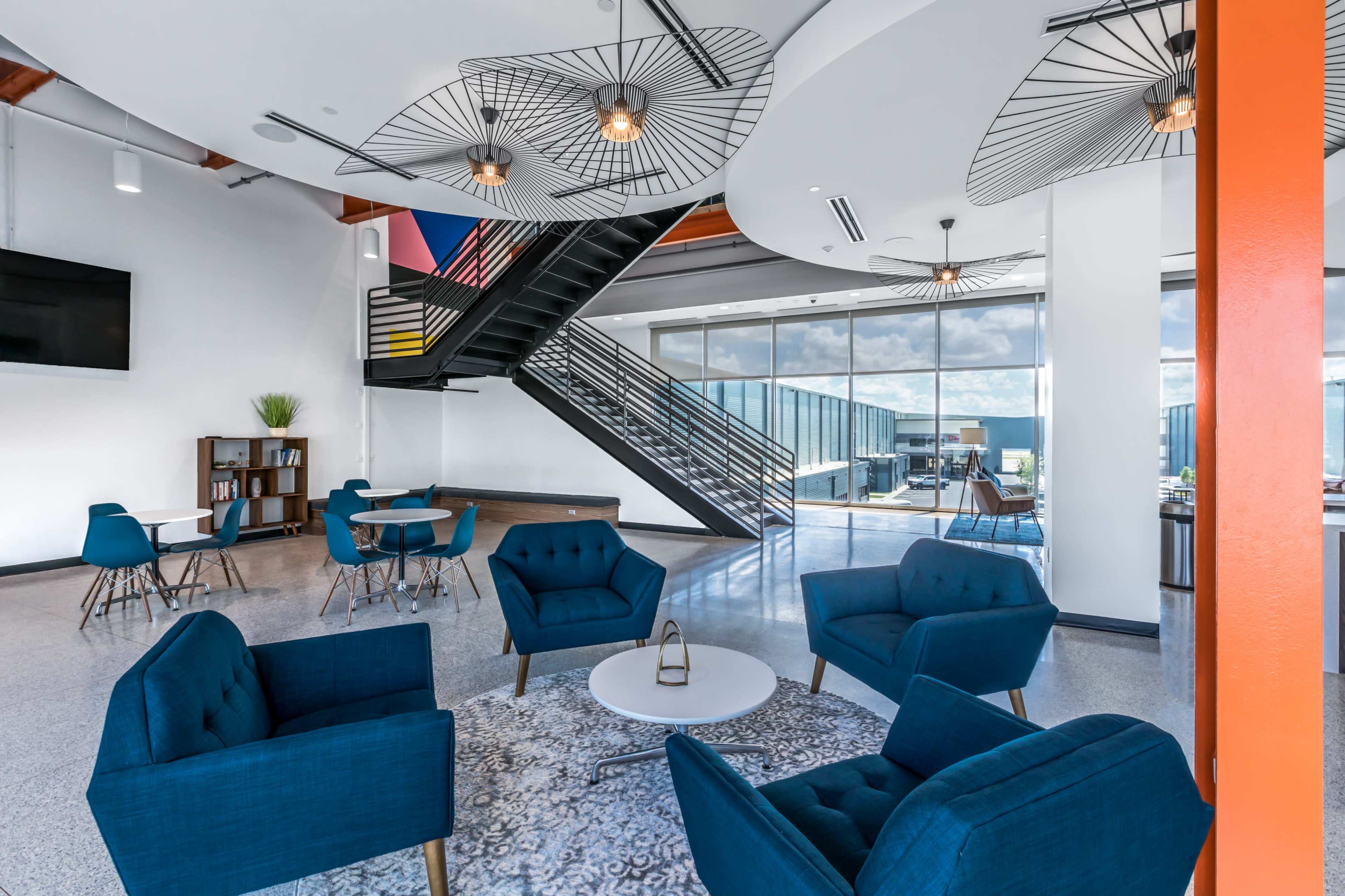 The image shows a modern lobby area with blue armchairs arranged around a white circular coffee table, a staircase in the background, and large windows allowing natural light to enter.