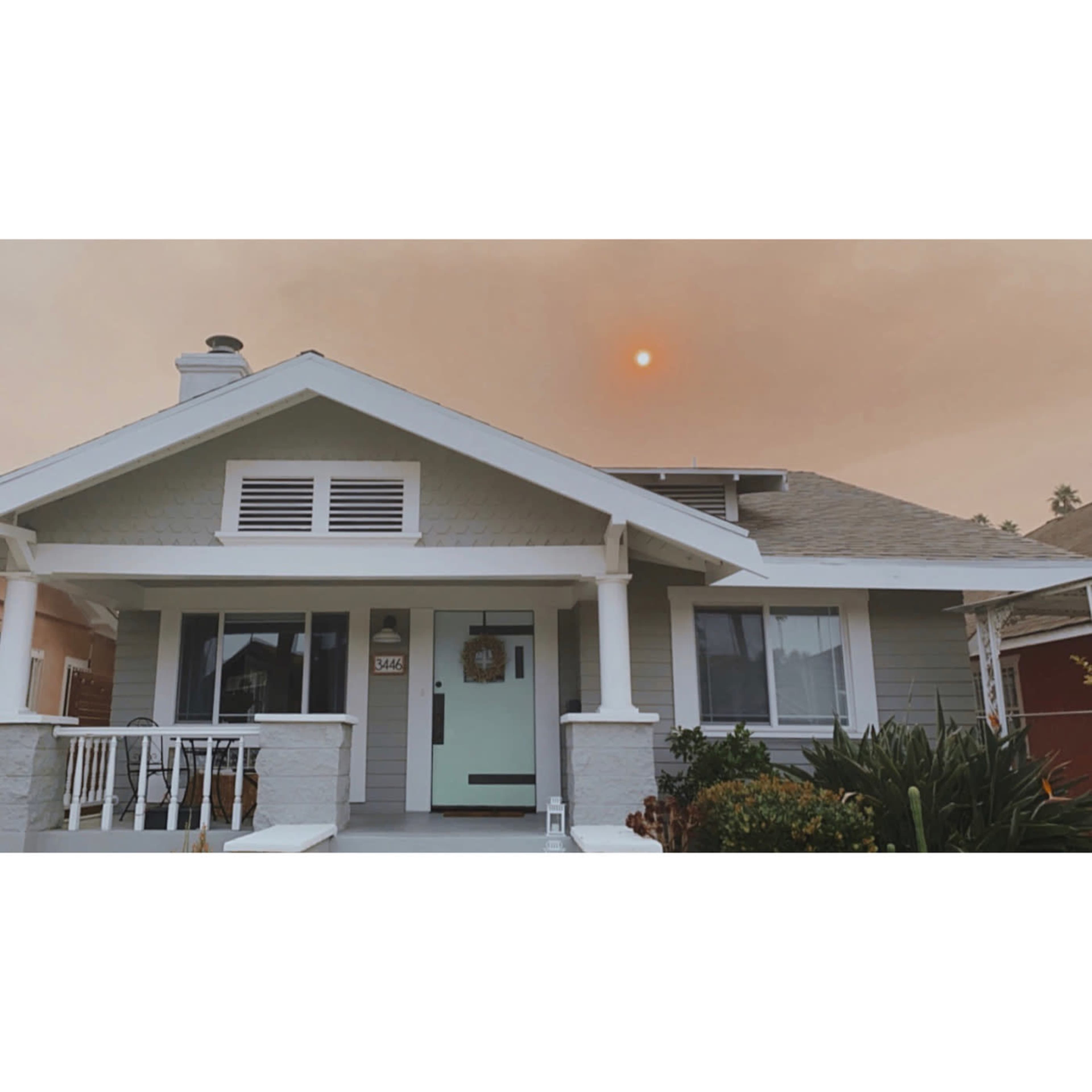 A single-story house with light gray siding and a mint green front door is shown under a hazy sky with a visible sun.