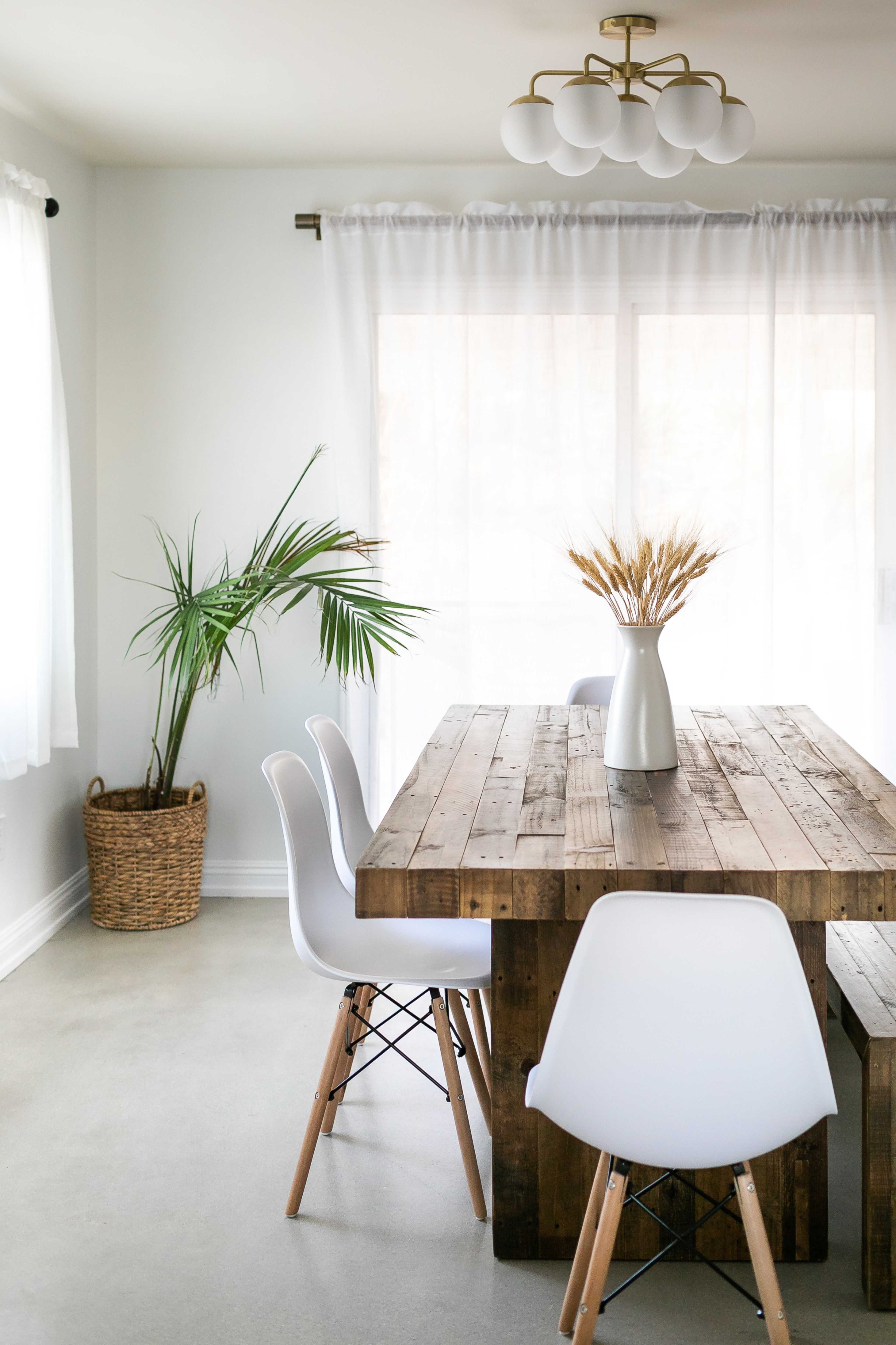 A modern dining area features a wooden table with a vase of dried plants, surrounded by white chairs and a potted palm in the corner.