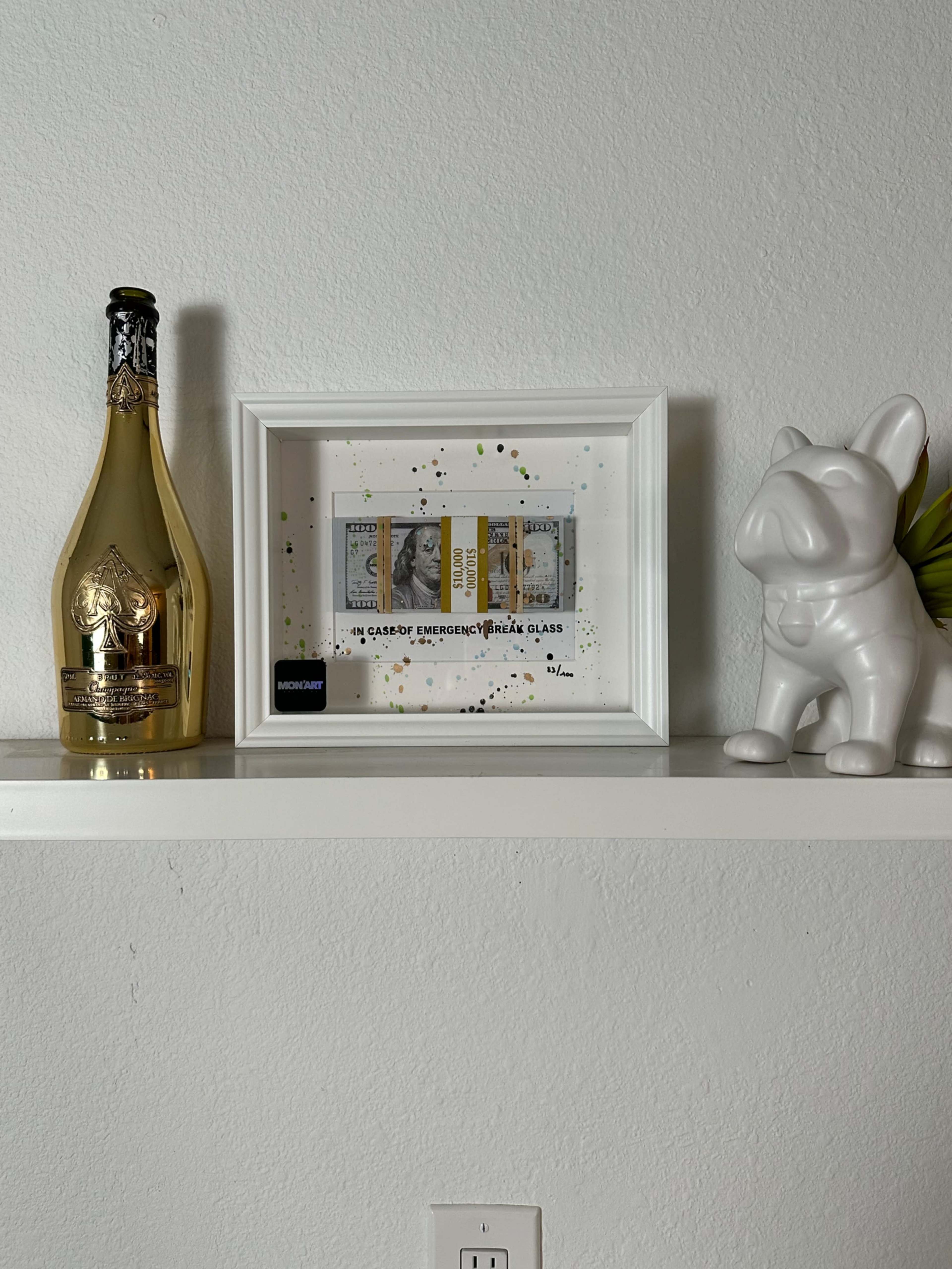 A white shelf displaying a framed set of banknotes, a gold bottle of champagne, and a white decorative dog figurine.