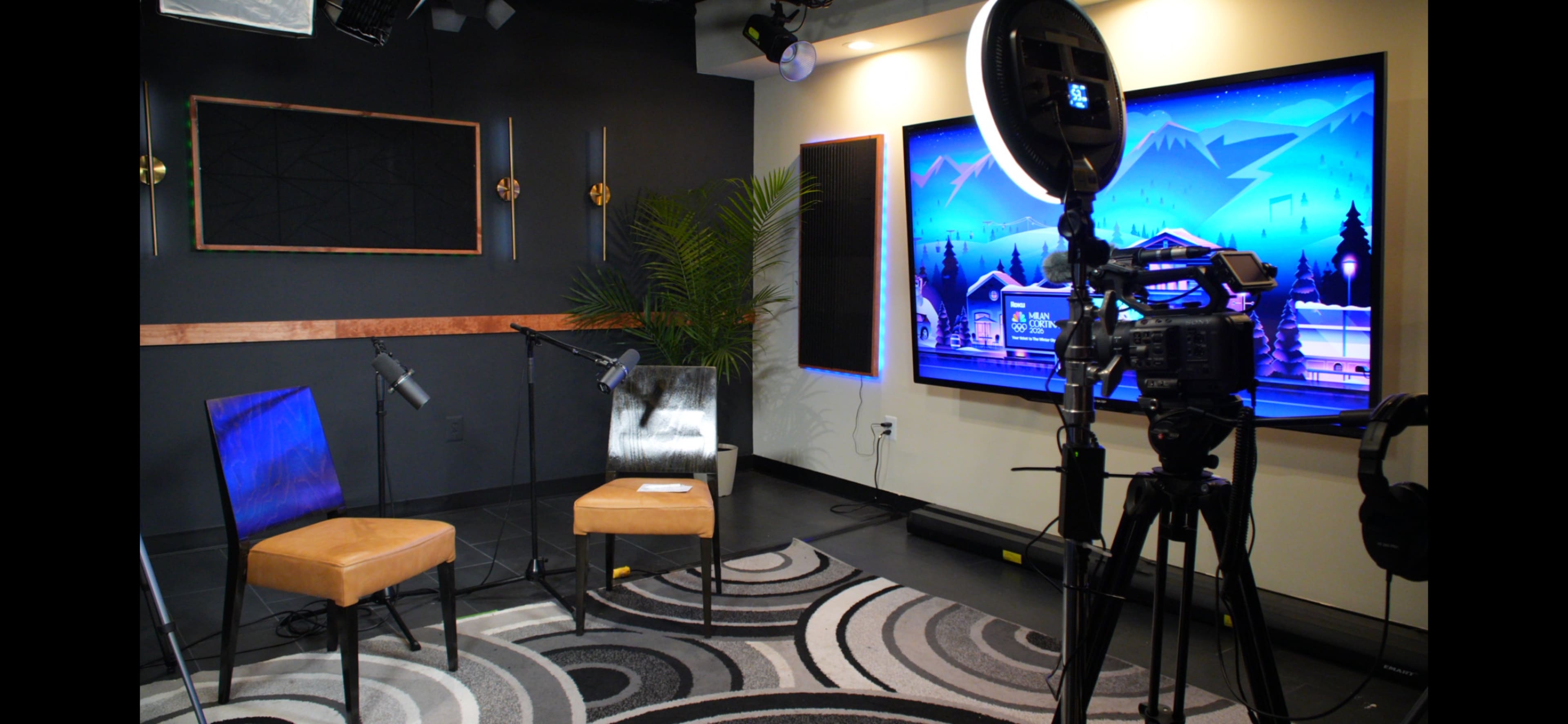 The image shows a television studio setup with two chairs, microphones, a camera on a tripod, and a large screen displaying a scenic backdrop.