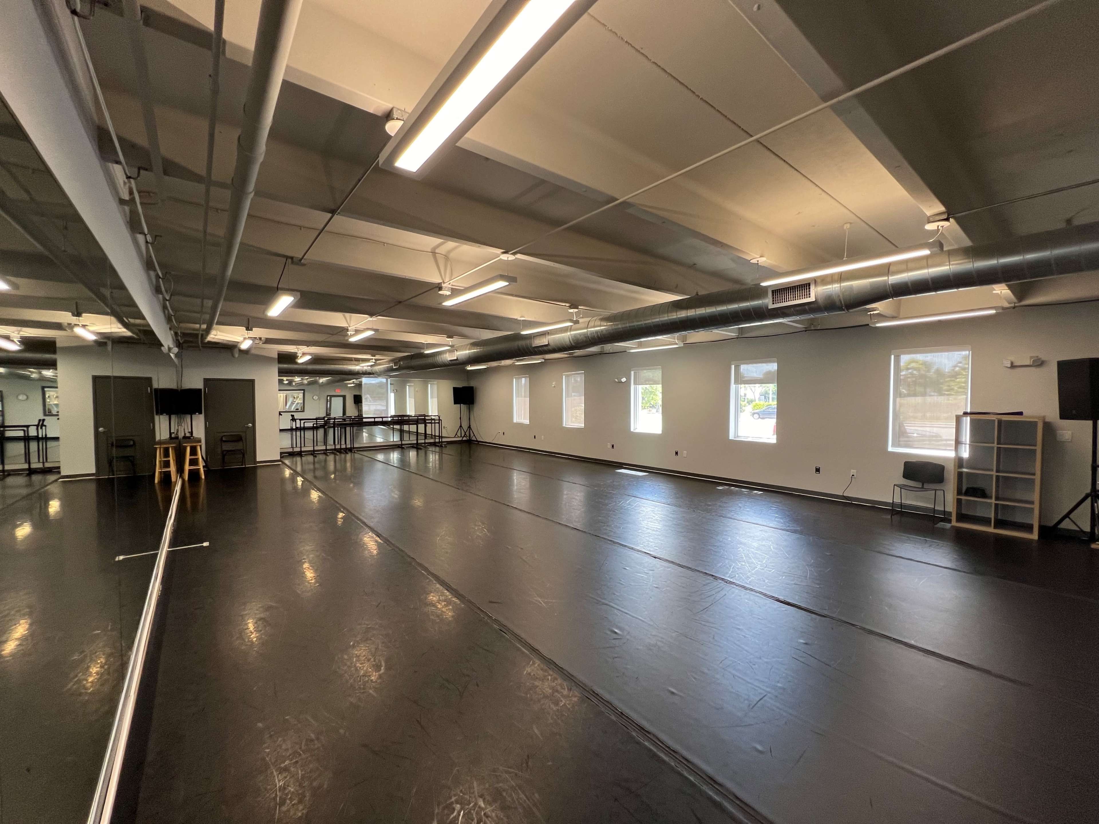 A spacious dance studio with a black floor, large mirrors, and several windows, equipped with speakers and a shelving unit.