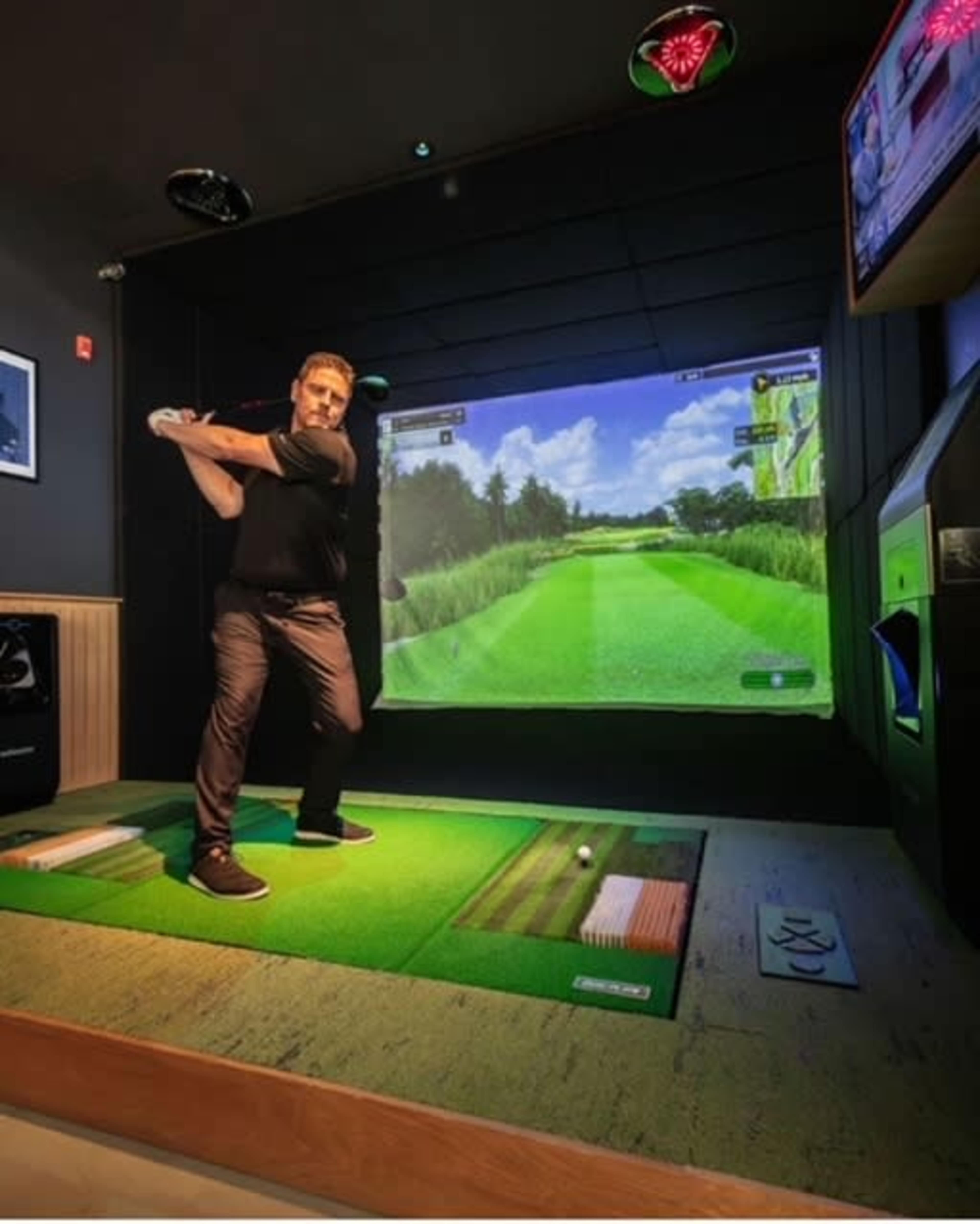 A man is swinging a golf club in front of a large screen showing a golf course simulation.