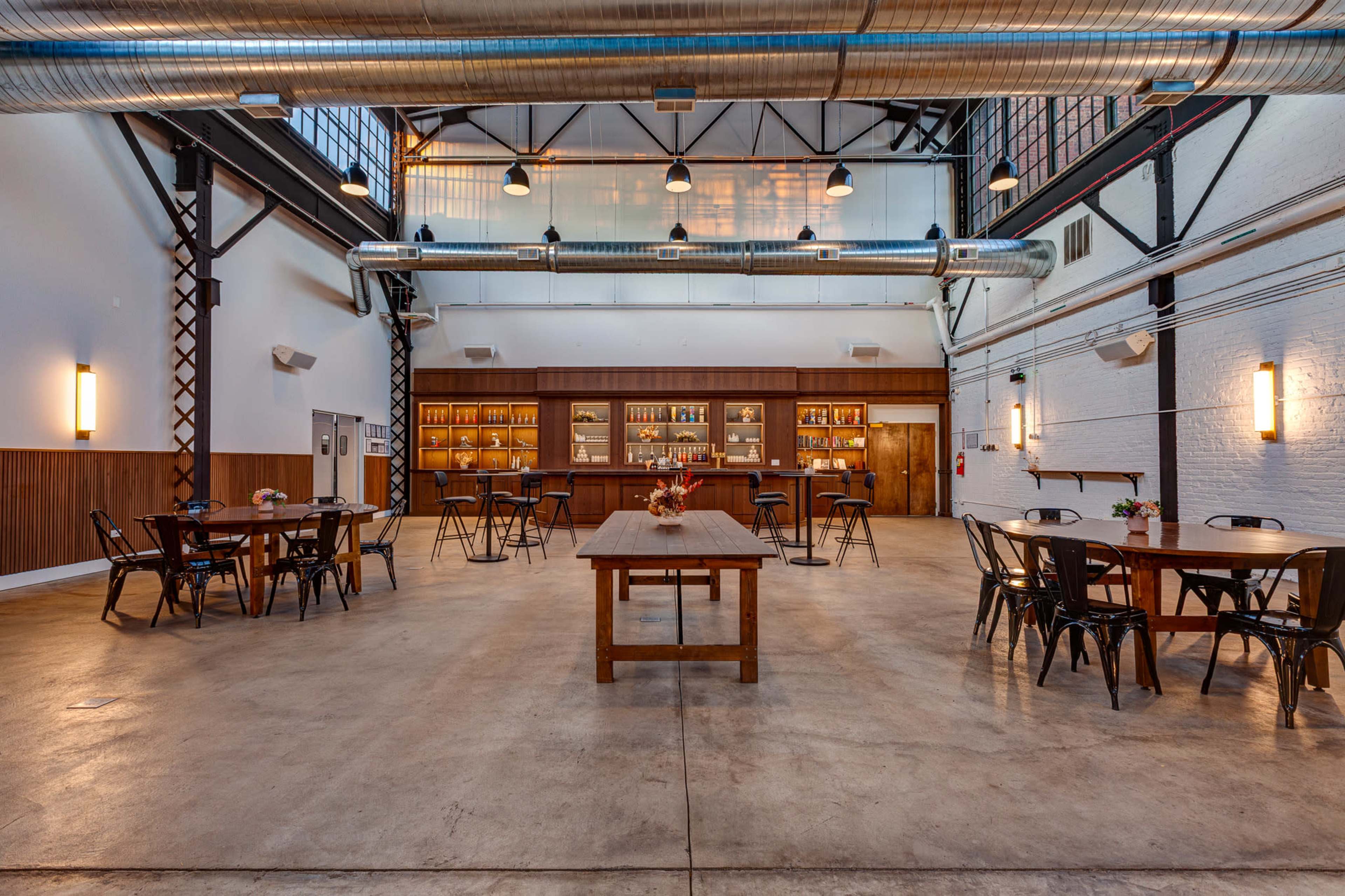 The image shows a spacious, industrial-style venue with wooden tables and chairs arranged around a central table, illuminated by overhead lights and featuring a bar along one wall.