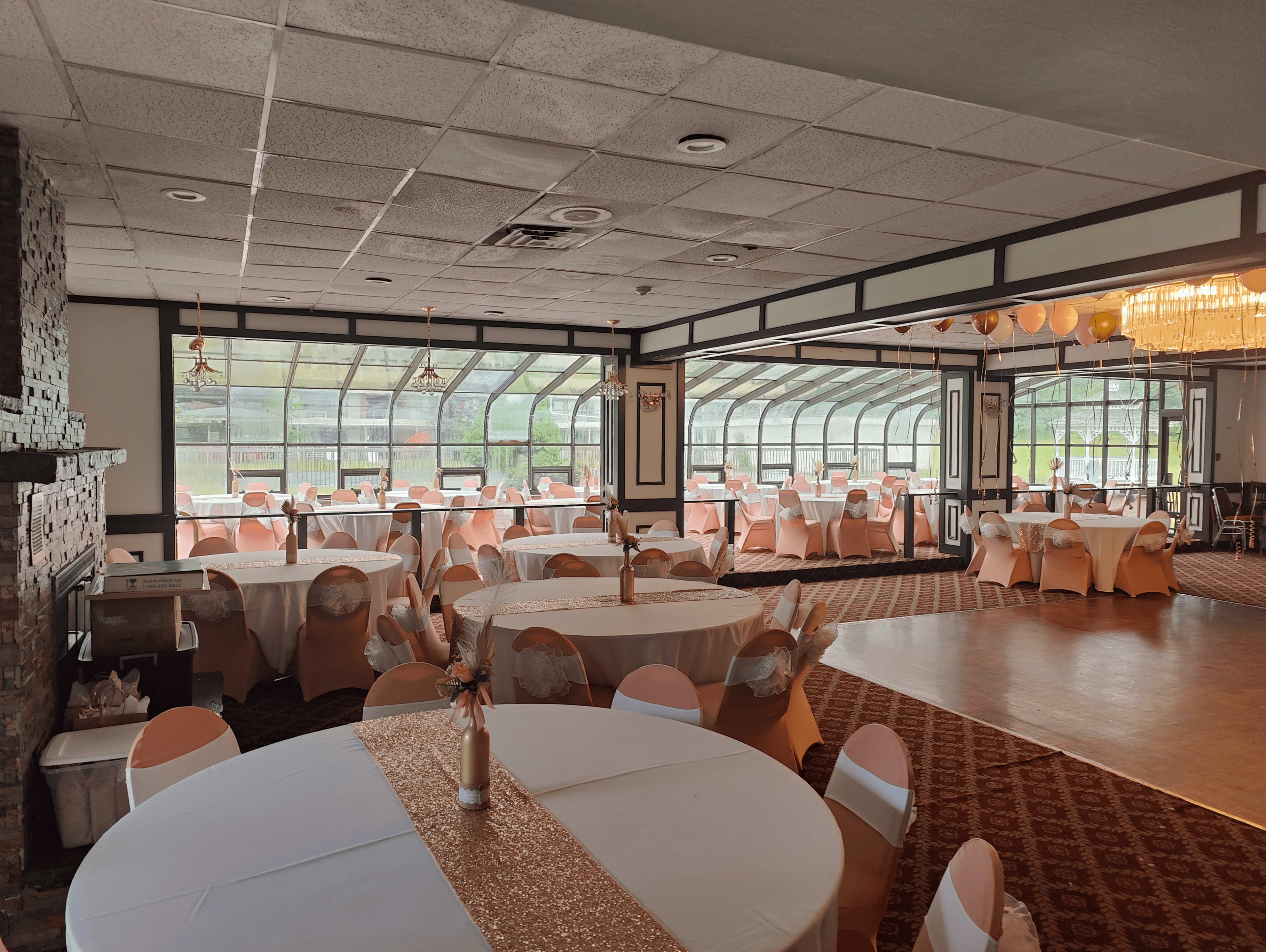 A spacious banquet hall with round tables covered in white tablecloths and adorned with peach-colored decorations, surrounded by large windows that provide ample natural light.