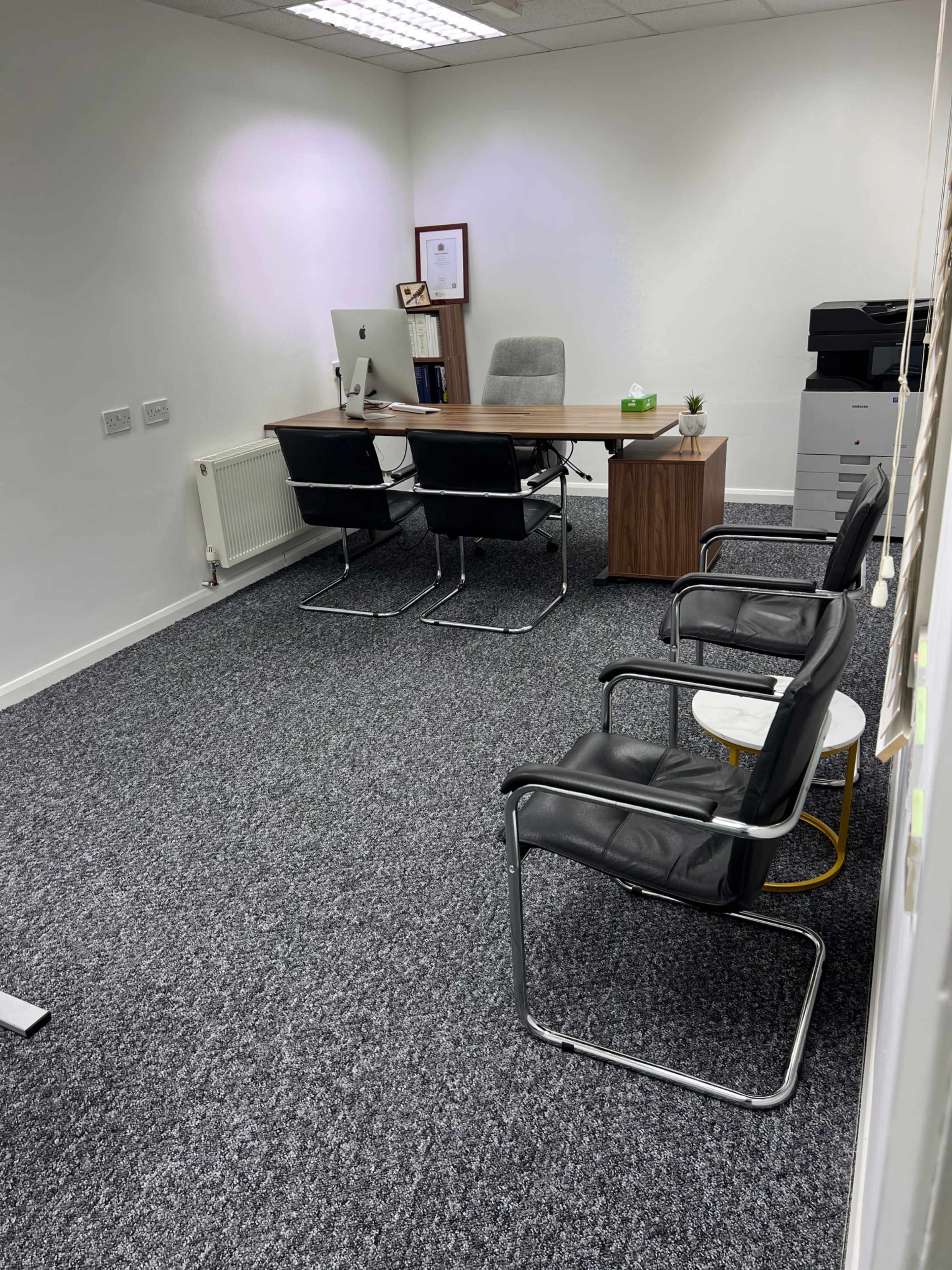 The image shows a minimalist office space featuring a wooden desk, an ergonomic chair, a desktop computer, and two black chairs arranged around a small round table.
