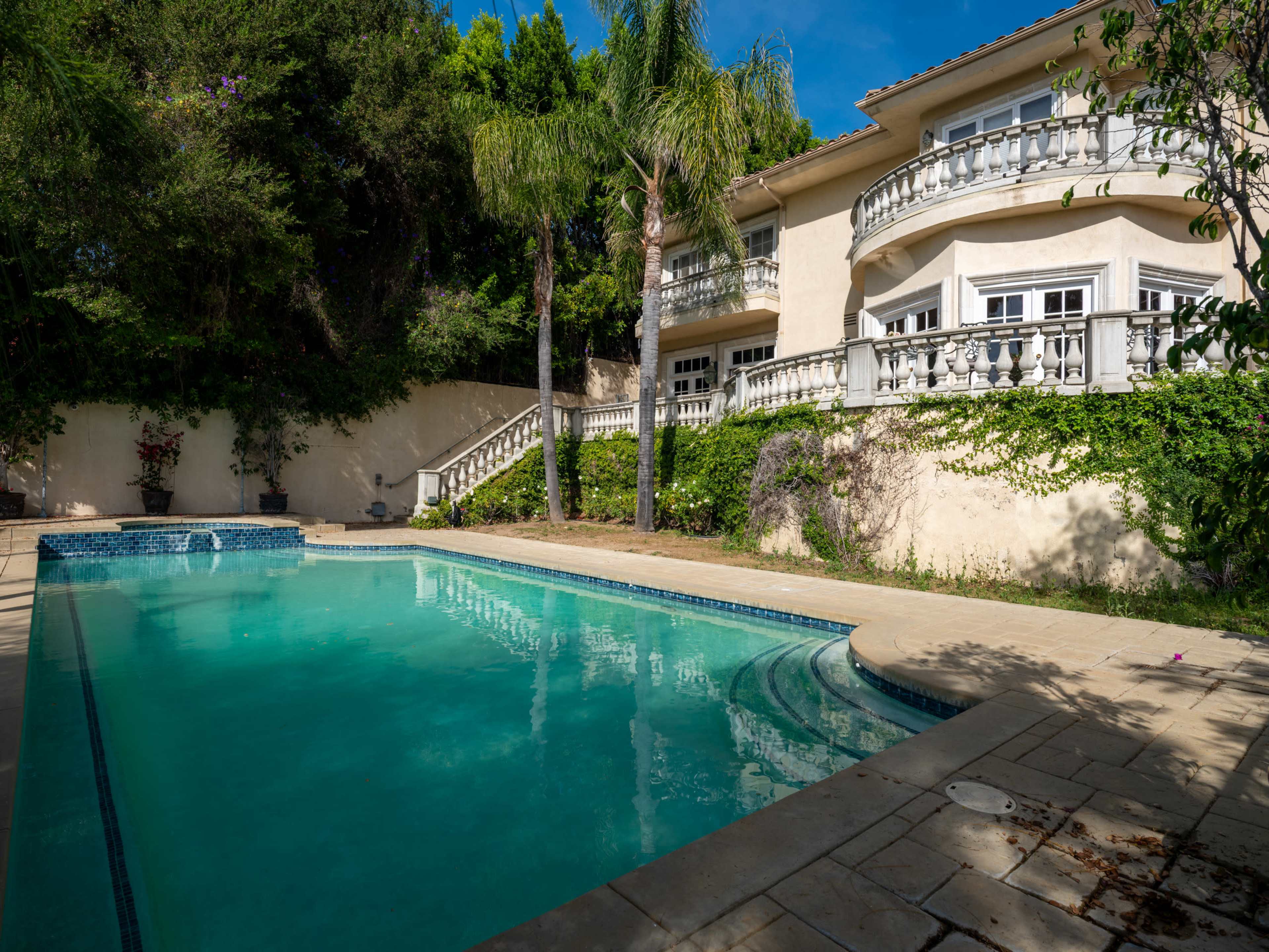 A clear swimming pool is situated in a backyard surrounded by lush greenery and a two-story house with balconies.