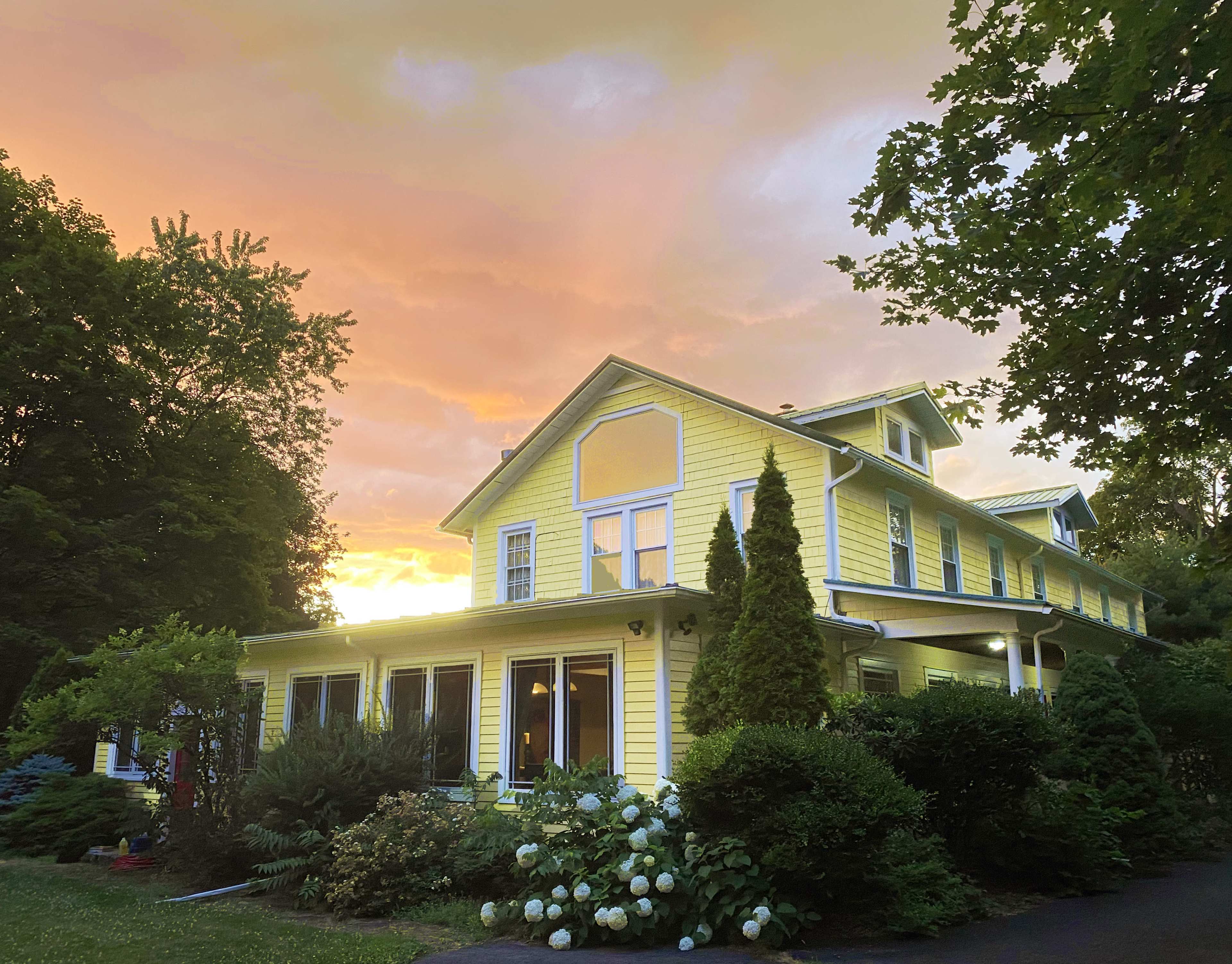 A yellow house with multiple windows is surrounded by trees and shrubs under a colorful sunset sky.