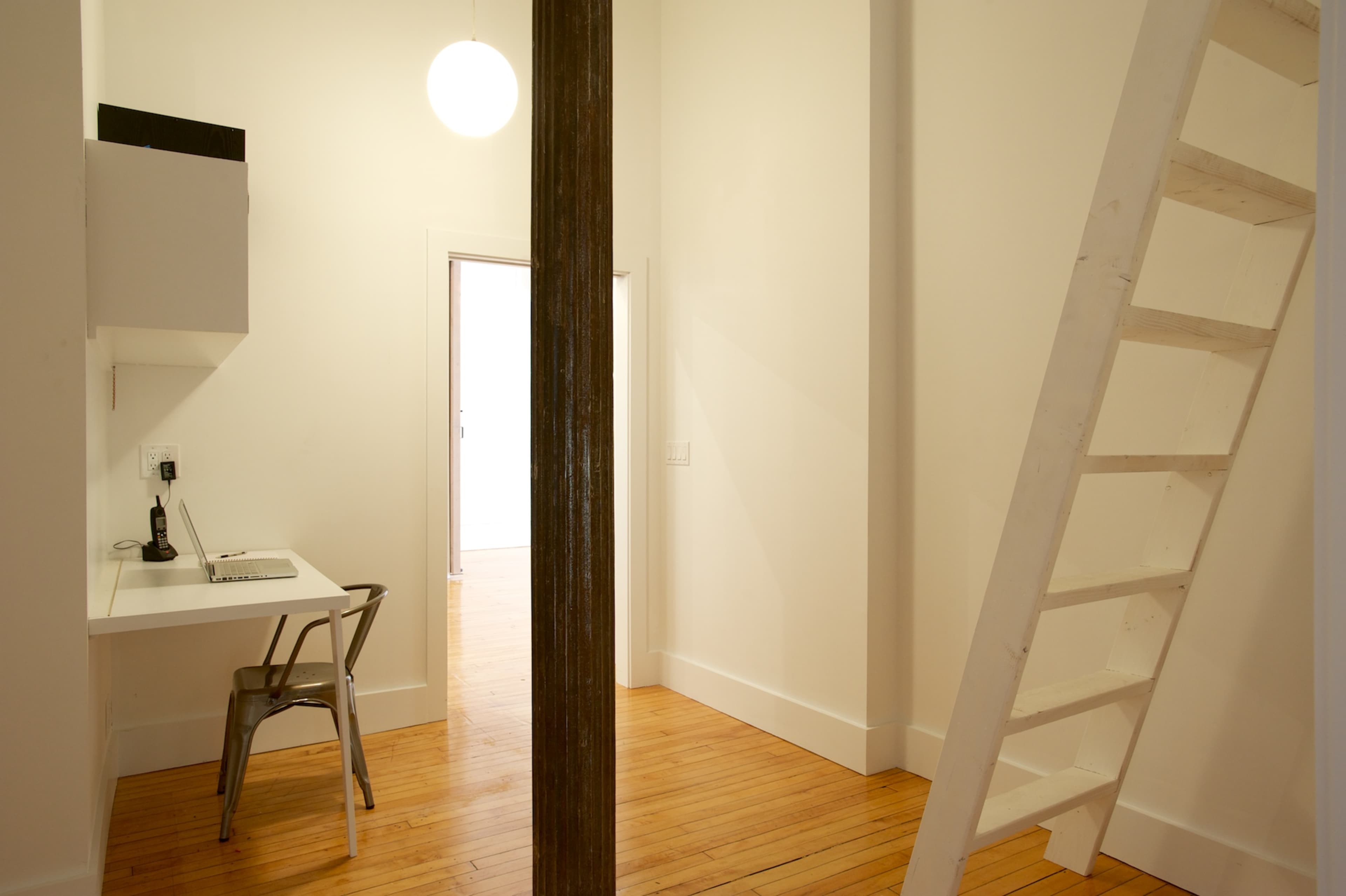 A minimalist room with a wooden floor, a small desk with a laptop, a metal chair, and a ladder leading to a loft area.