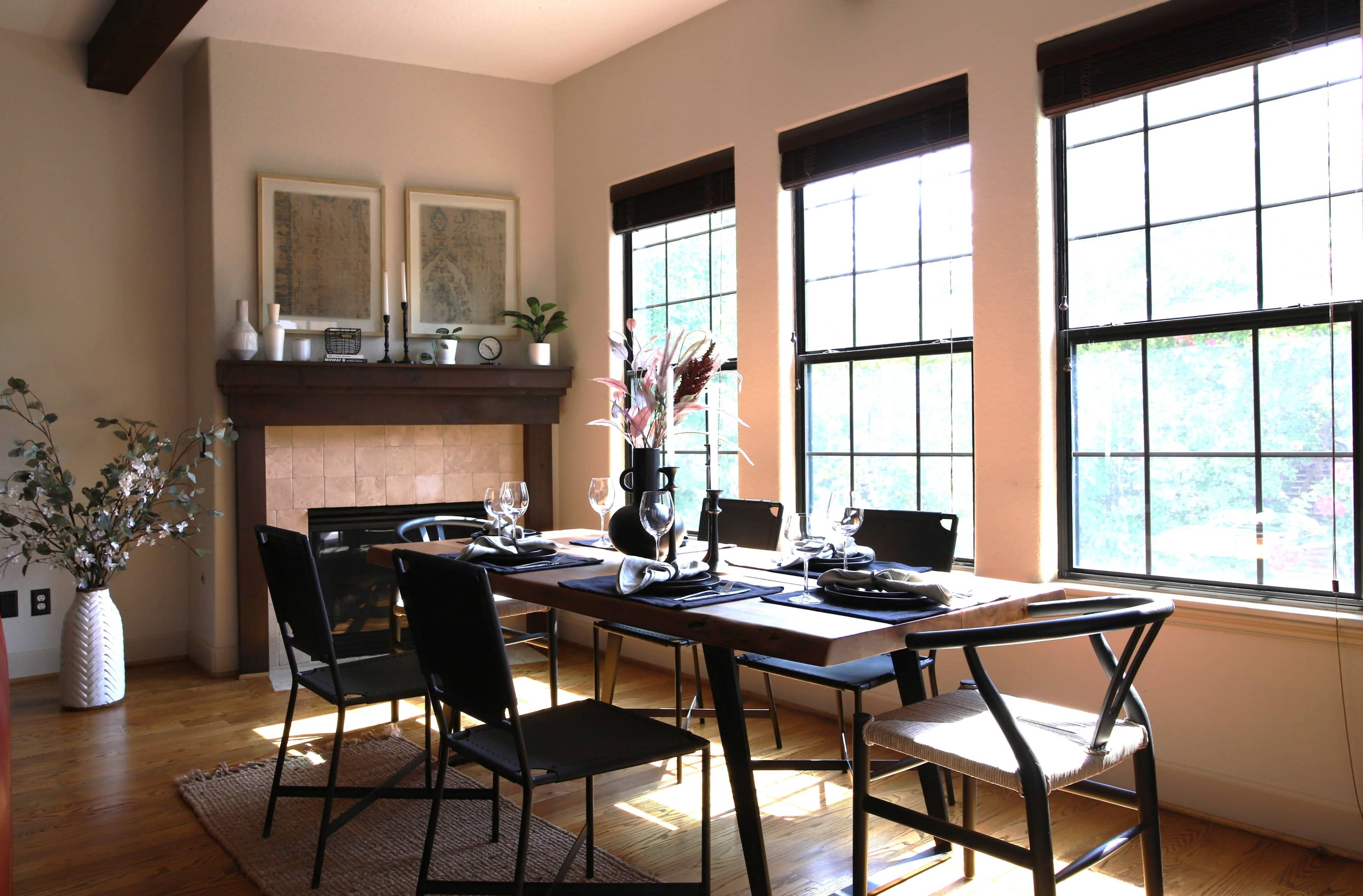 A dining room features a wooden table set for a meal, surrounded by black chairs, with large windows allowing natural light and a fireplace along one wall.