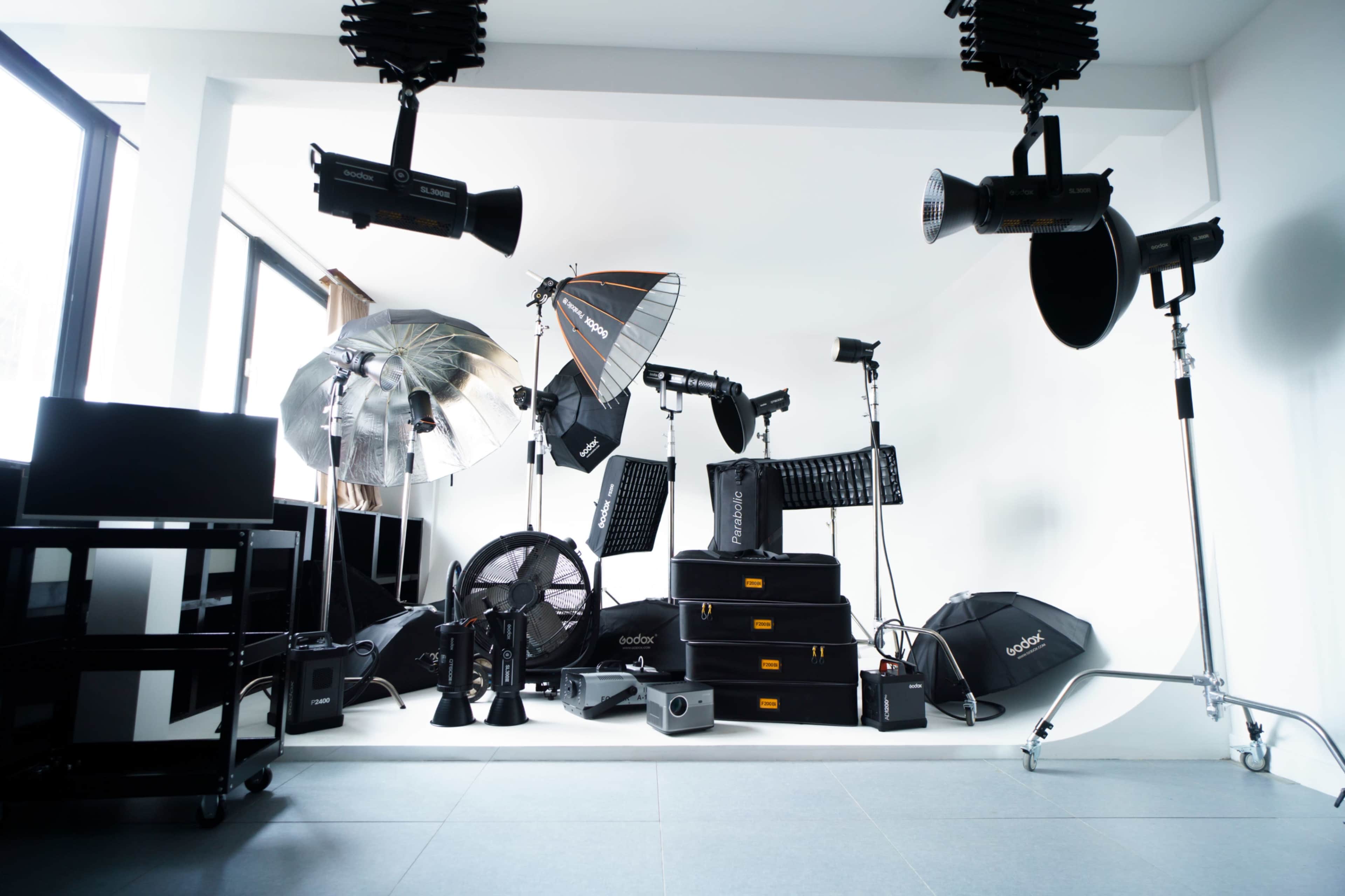The image shows a photography studio equipped with various lighting equipment, including softboxes, reflectors, and stands, arranged on a clean, minimalistic backdrop.