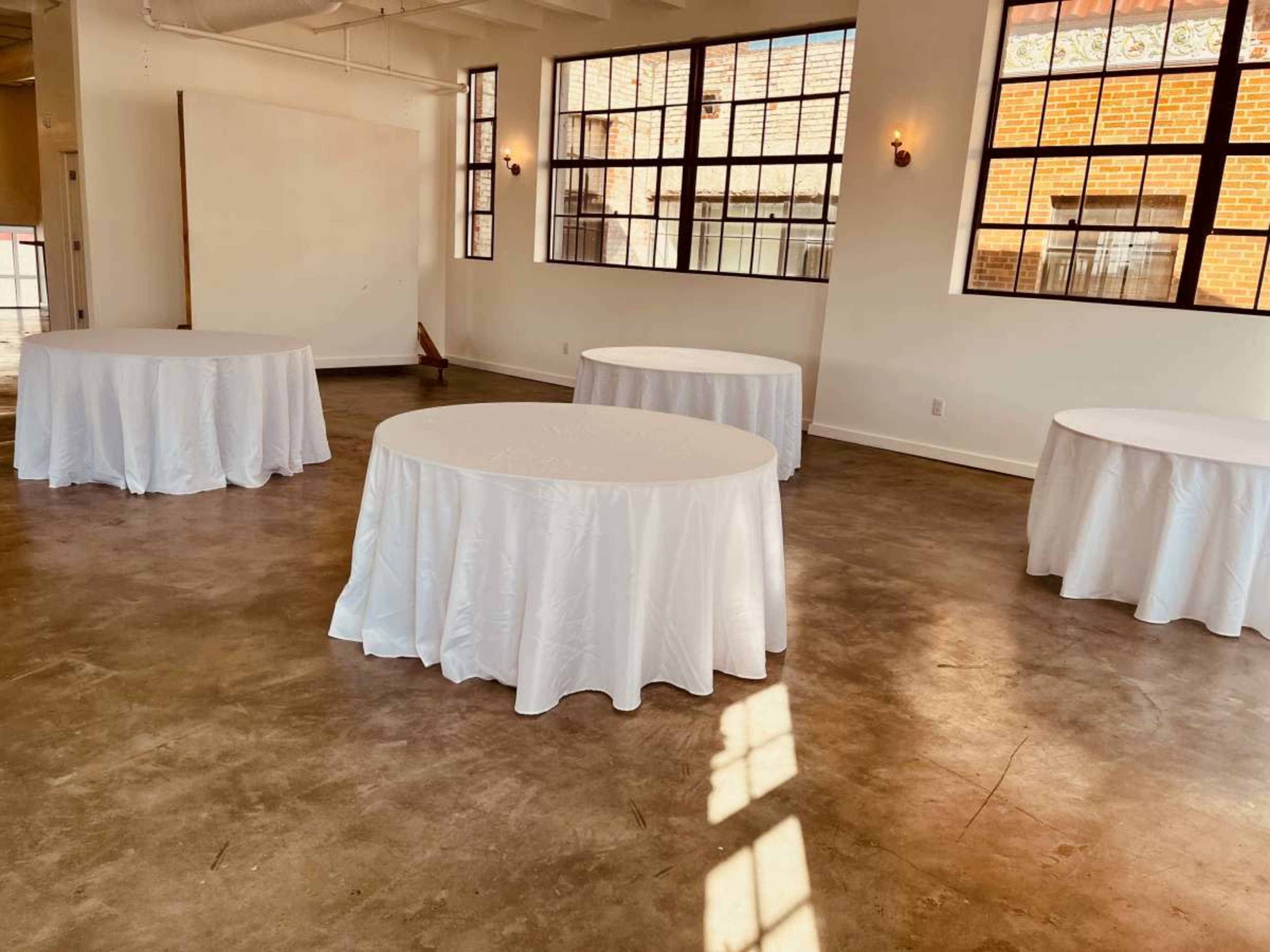 The image shows a bright room with large windows featuring four round tables covered with white tablecloths on a concrete floor.