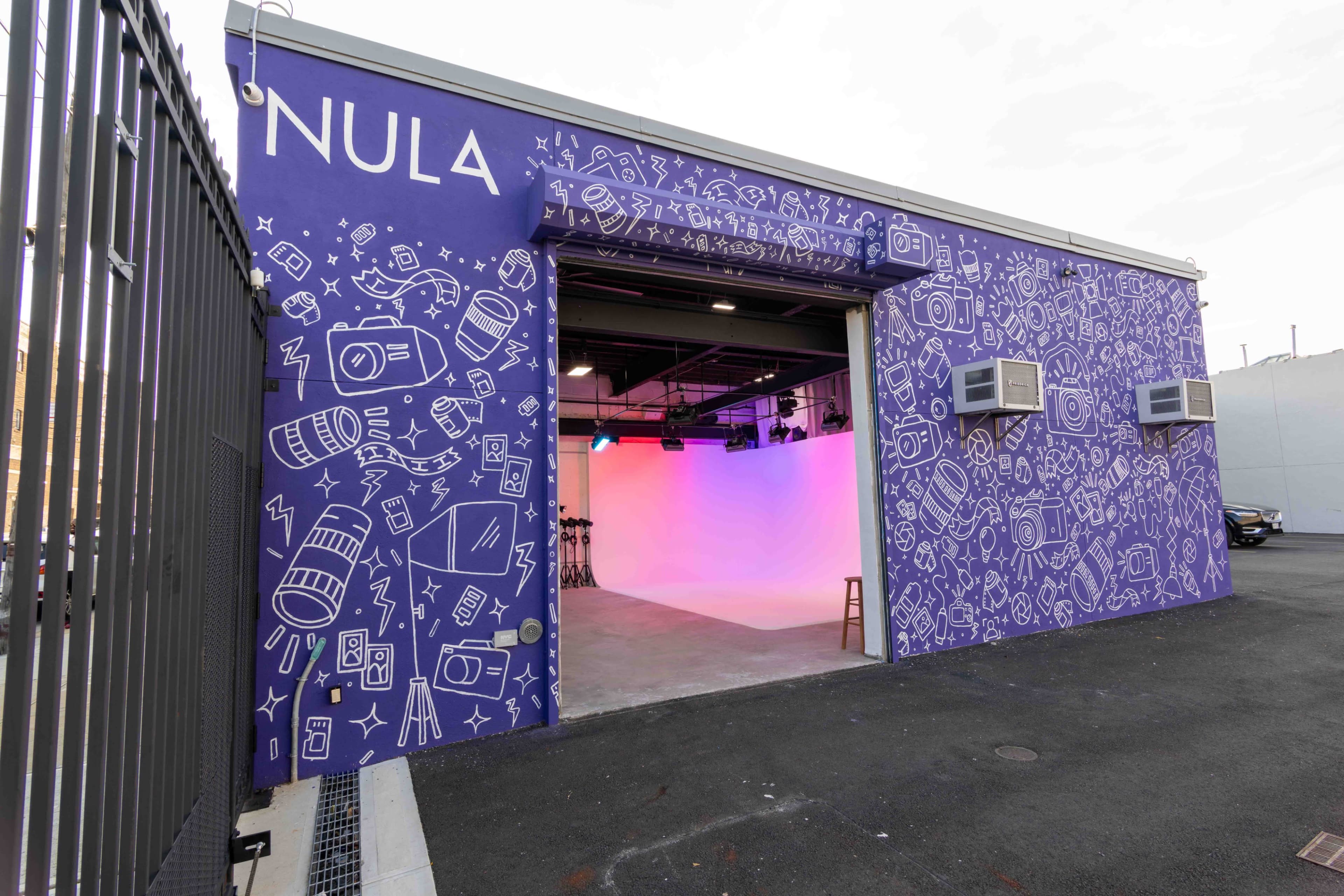 A purple building with an open garage door features a white studio space illuminated with colorful lighting and decorated with graphic camera and film illustrations on the exterior walls.