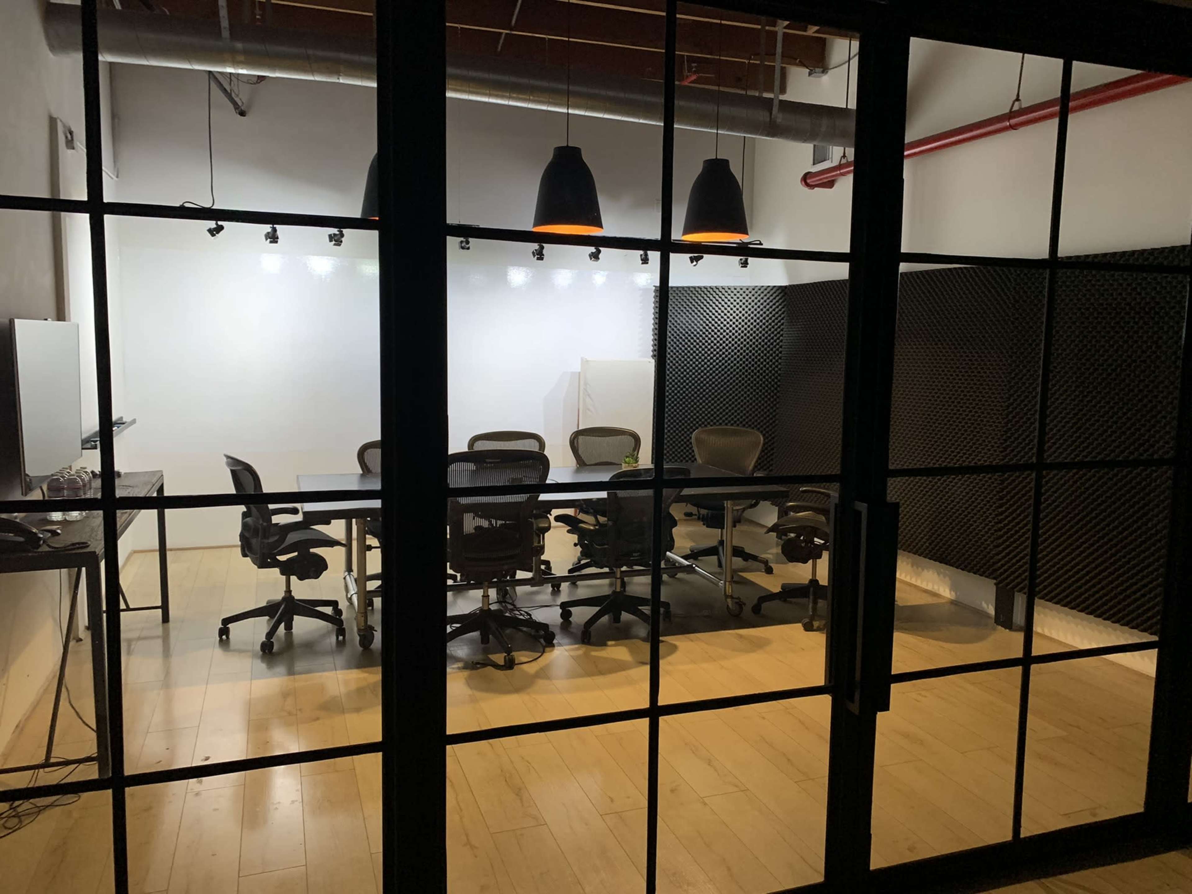 A conference room with a large table and several office chairs is visible through glass doors, featuring acoustic paneling on the walls and overhead lighting.