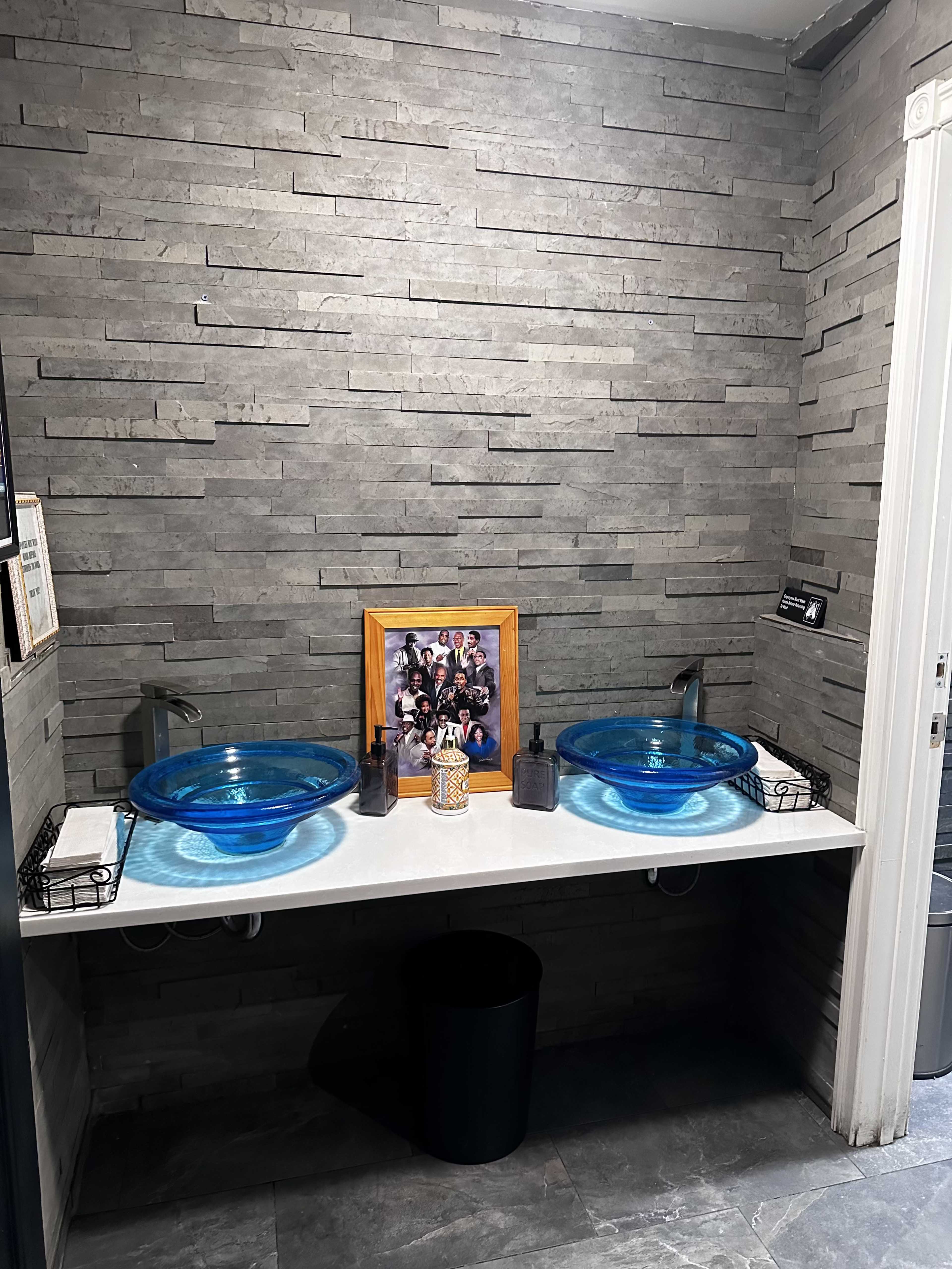 The image shows a bathroom with two blue glass sinks on a white countertop, set against a textured gray stone wall, with a framed photo and various toiletries in the center.