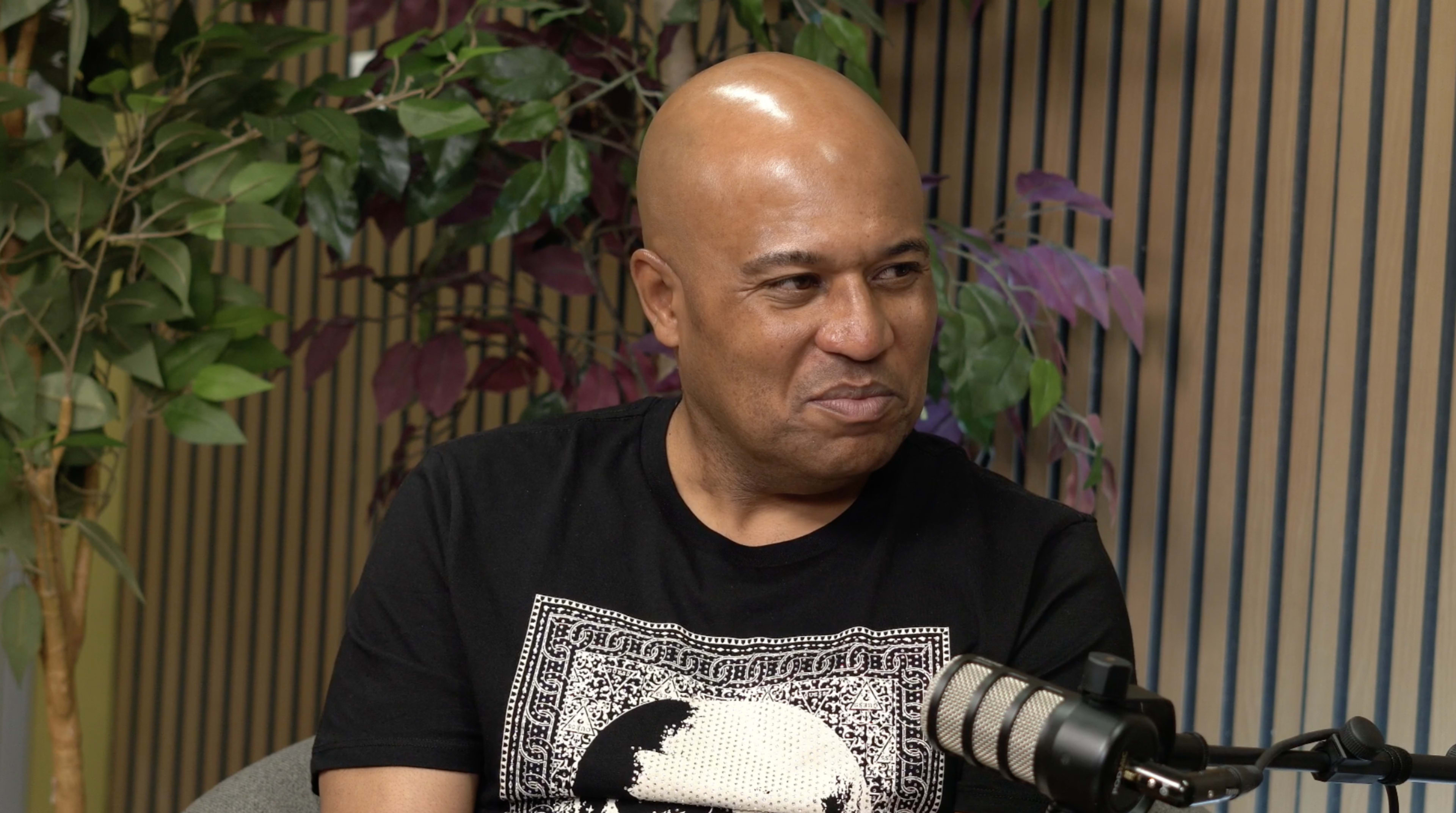 A bald man in a black T-shirt sits in front of a microphone, surrounded by greenery and a striped backdrop.