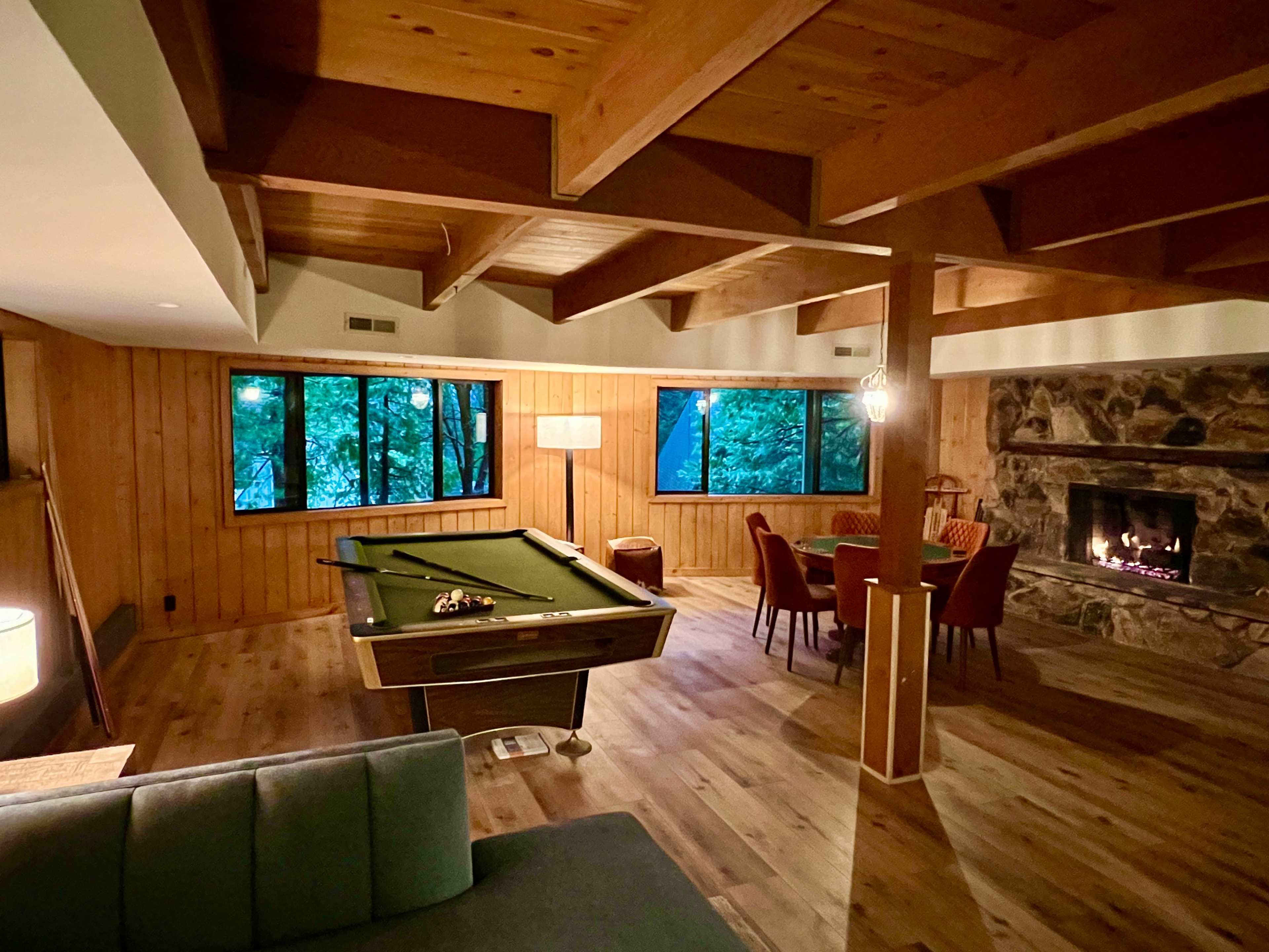 A spacious, wooden-paneled living room features a billiard table, a dining area, and large windows overlooking a wooded landscape.