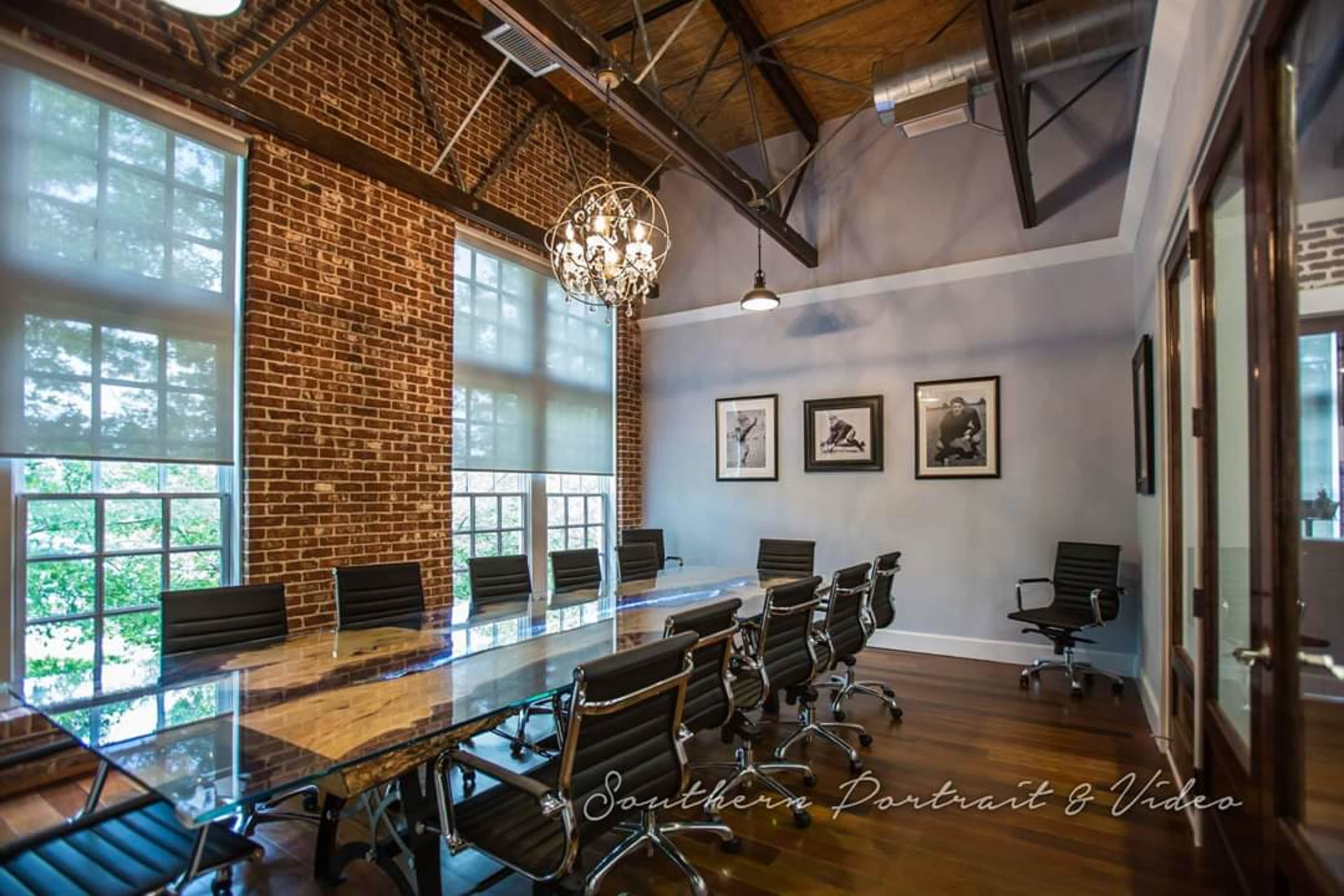 A modern conference room featuring a long glass table, black office chairs, exposed brick walls, and framed artwork on the walls.