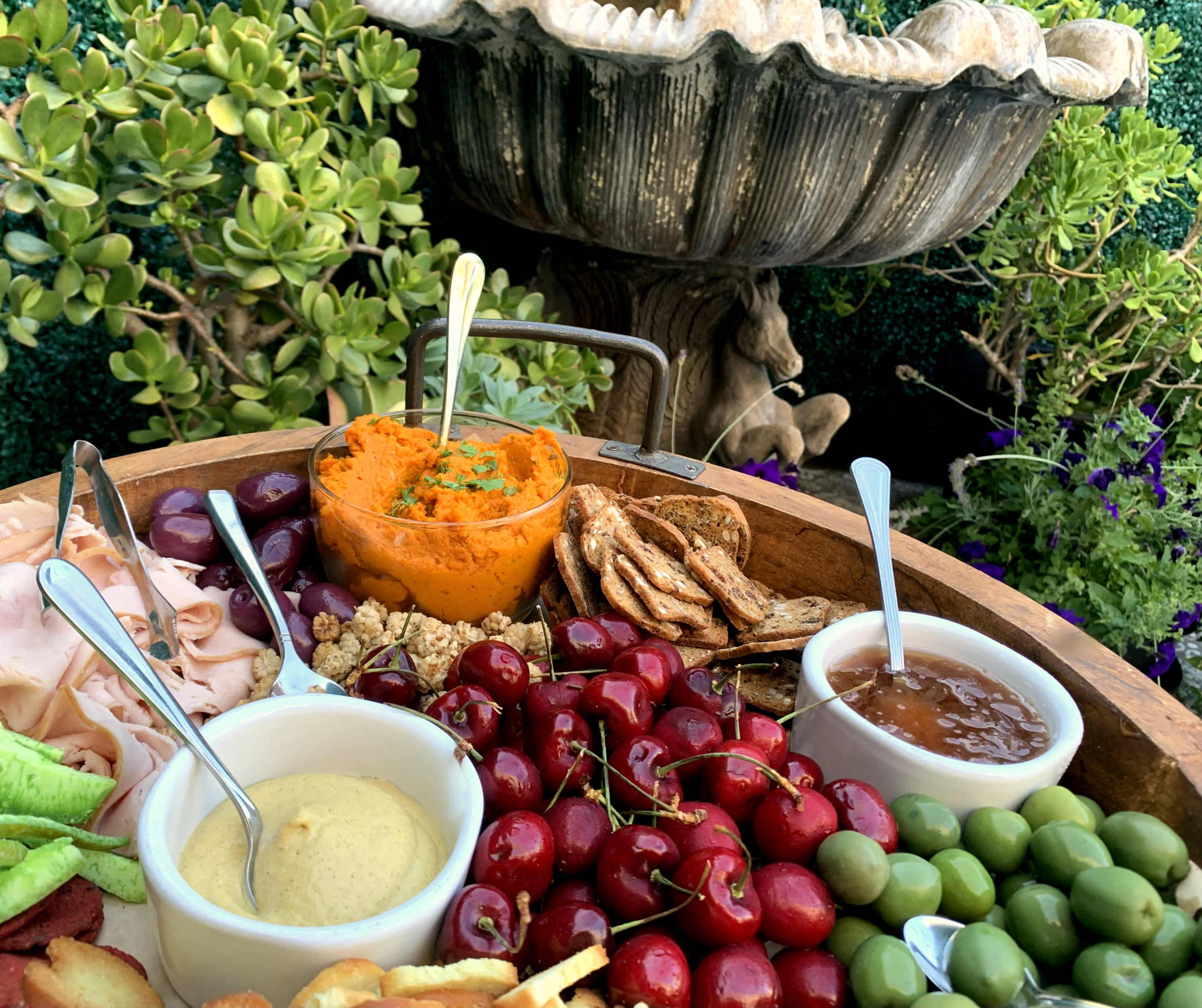 A wooden platter filled with a variety of meats, cheeses, fruits, and dips is set in front of a decorative shell-shaped fountain surrounded by greenery.