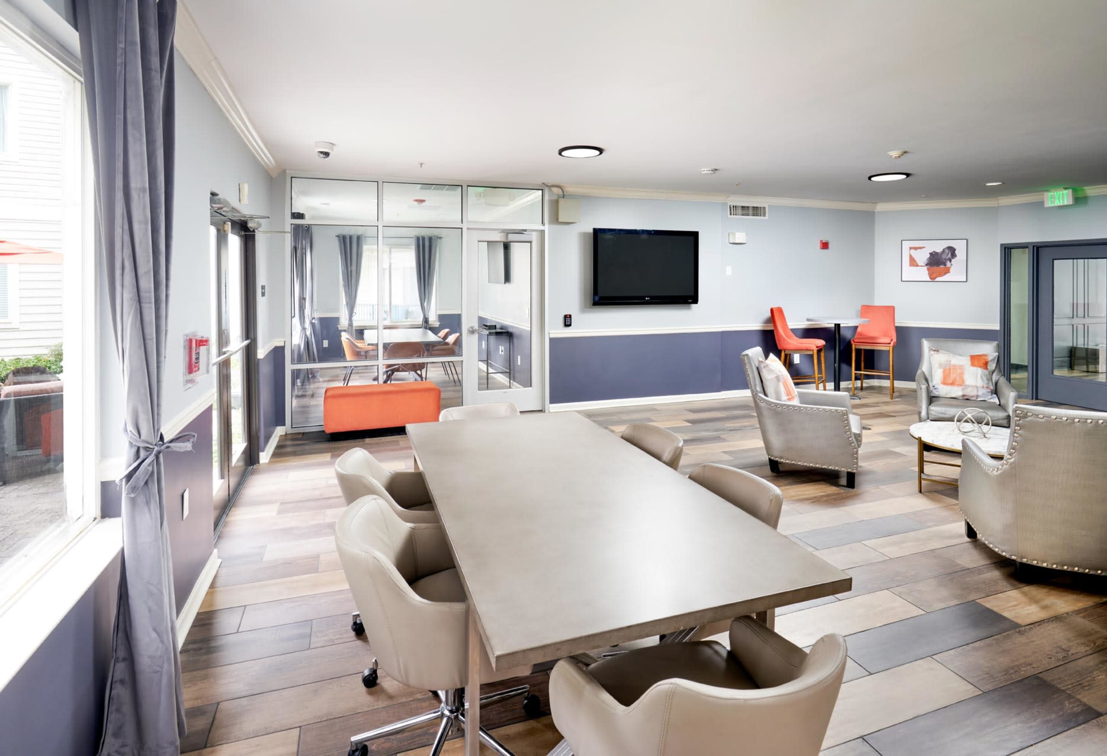 The image shows a modern lounge area featuring a large table with chairs, a television mounted on the wall, and various seating arrangements around the space.