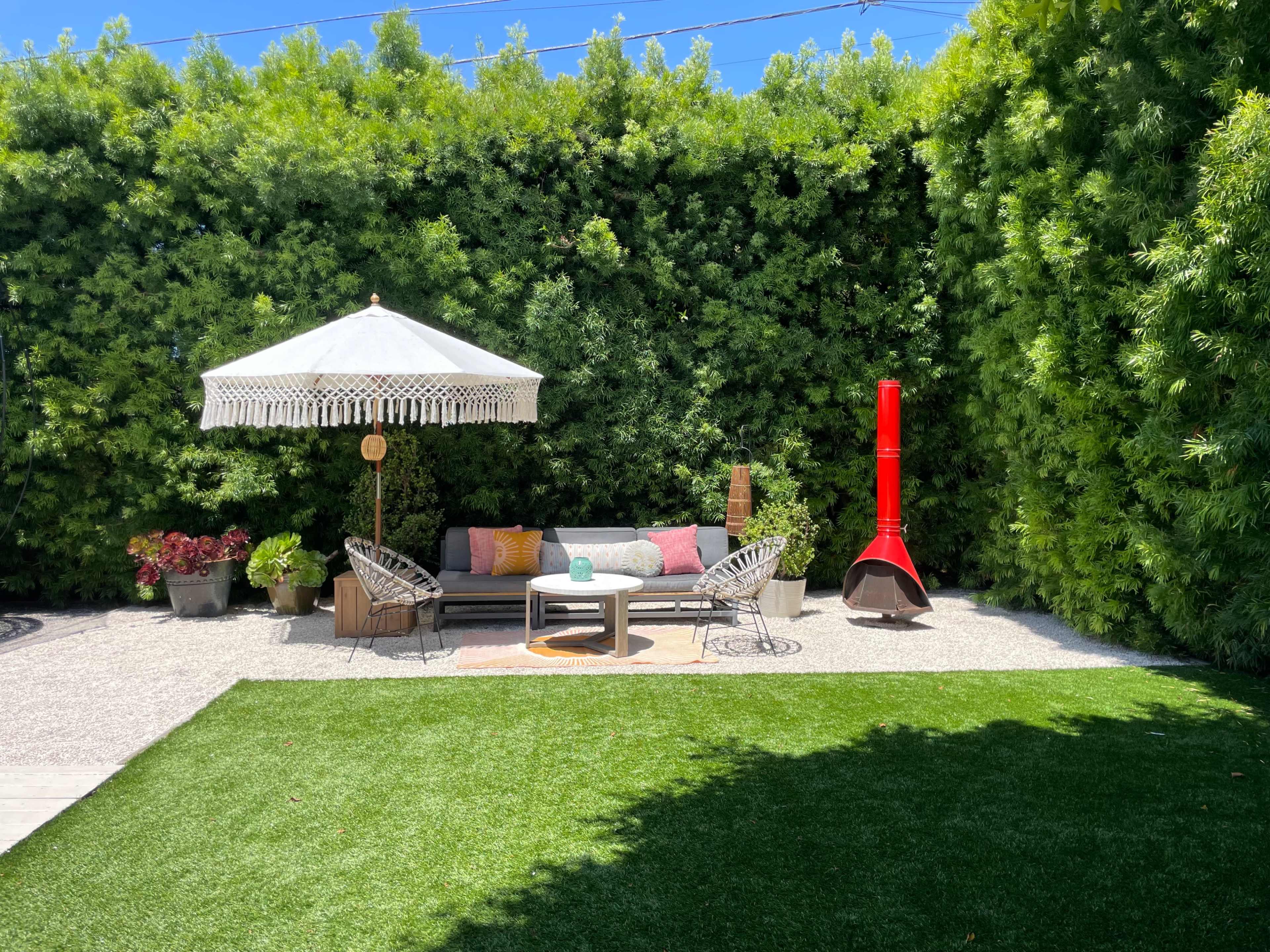 A cozy outdoor seating area features a shaded umbrella, a fire pit, and planters, surrounded by lush greenery.