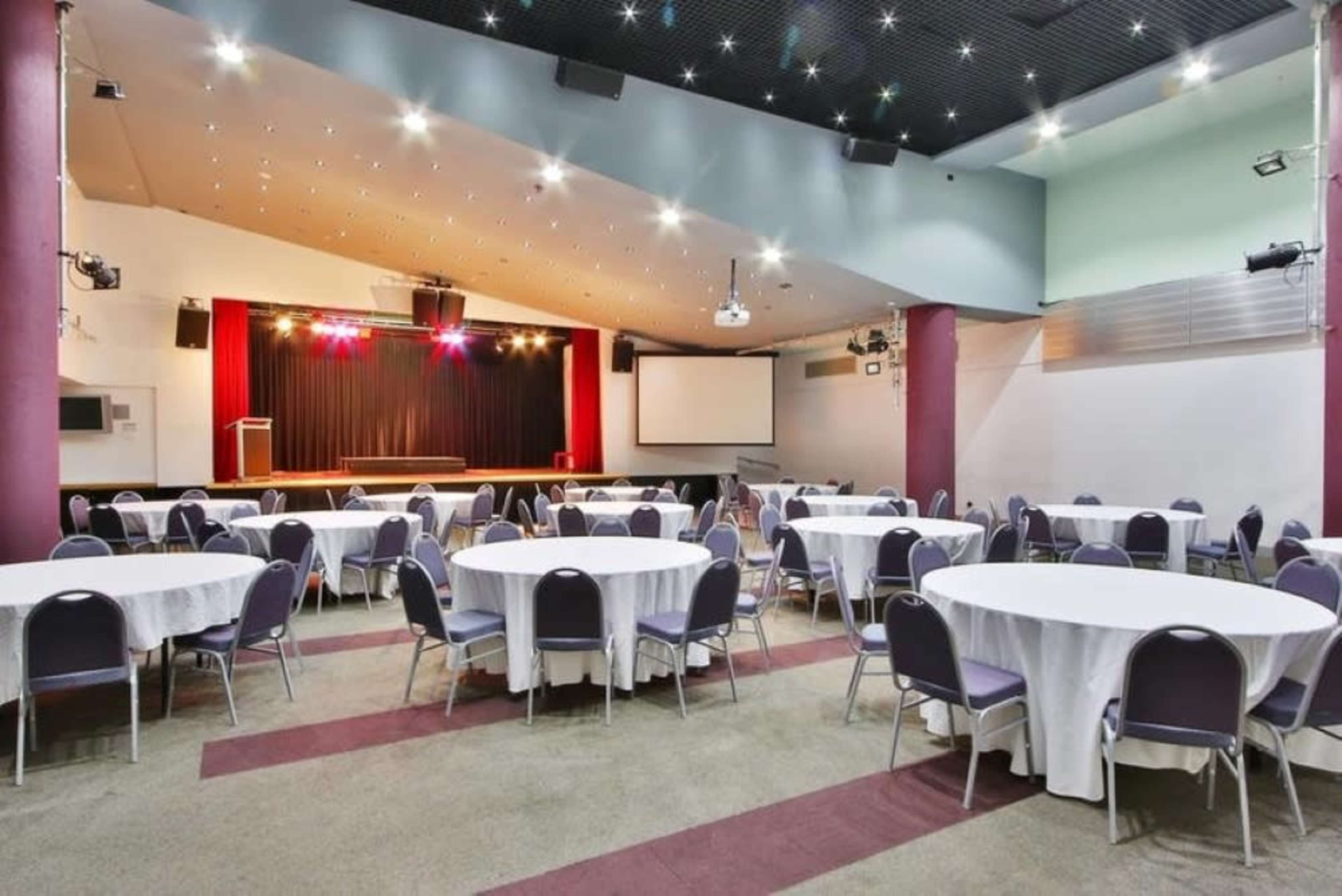 The image shows a spacious event hall with round tables arranged in rows and a stage at the front, illuminated by stage lights.