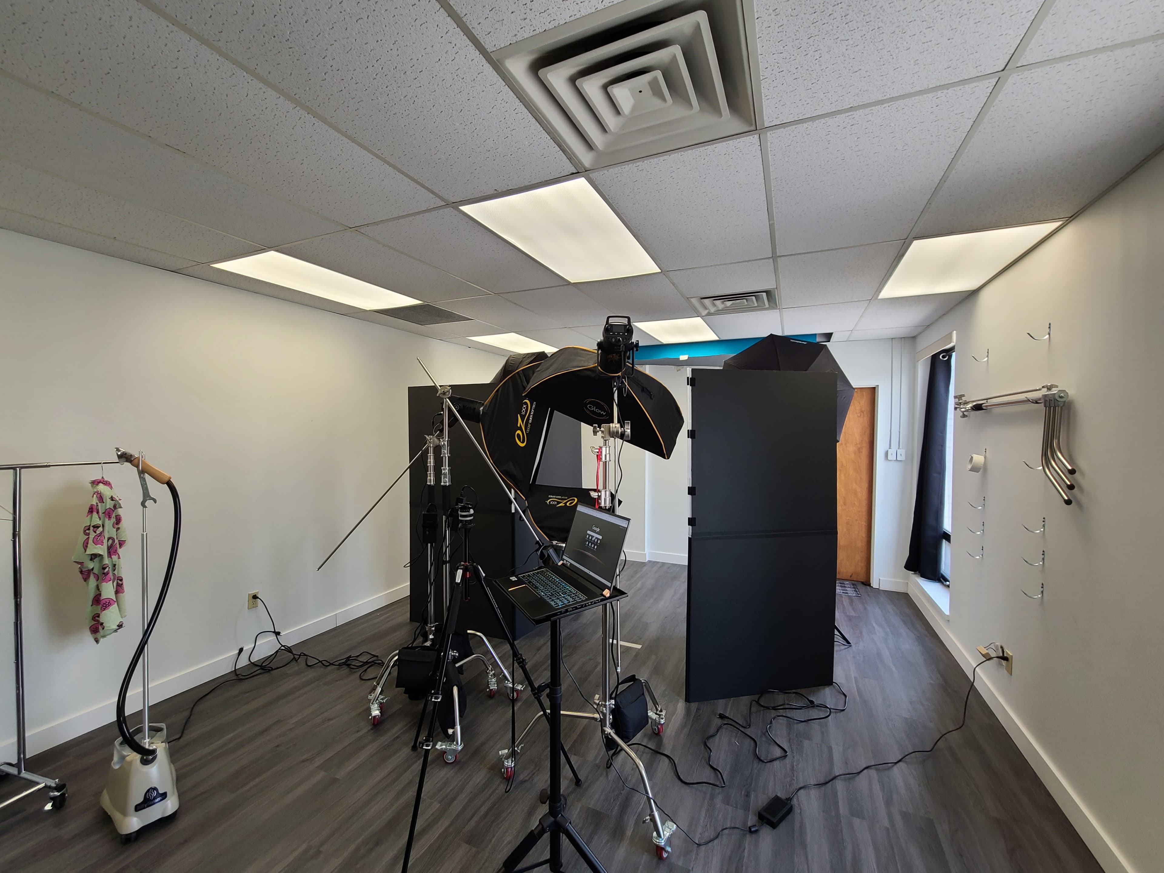 A photography studio setup with lighting equipment, a laptop, and dark backdrops in a spacious room.
